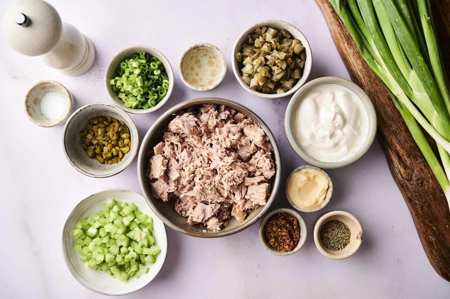 A top-down shot of the ingredients for a dill pickle tuna salad, including a bowl of tuna, chopped celery and pickles, and small bowls of mayonnaise and spices.