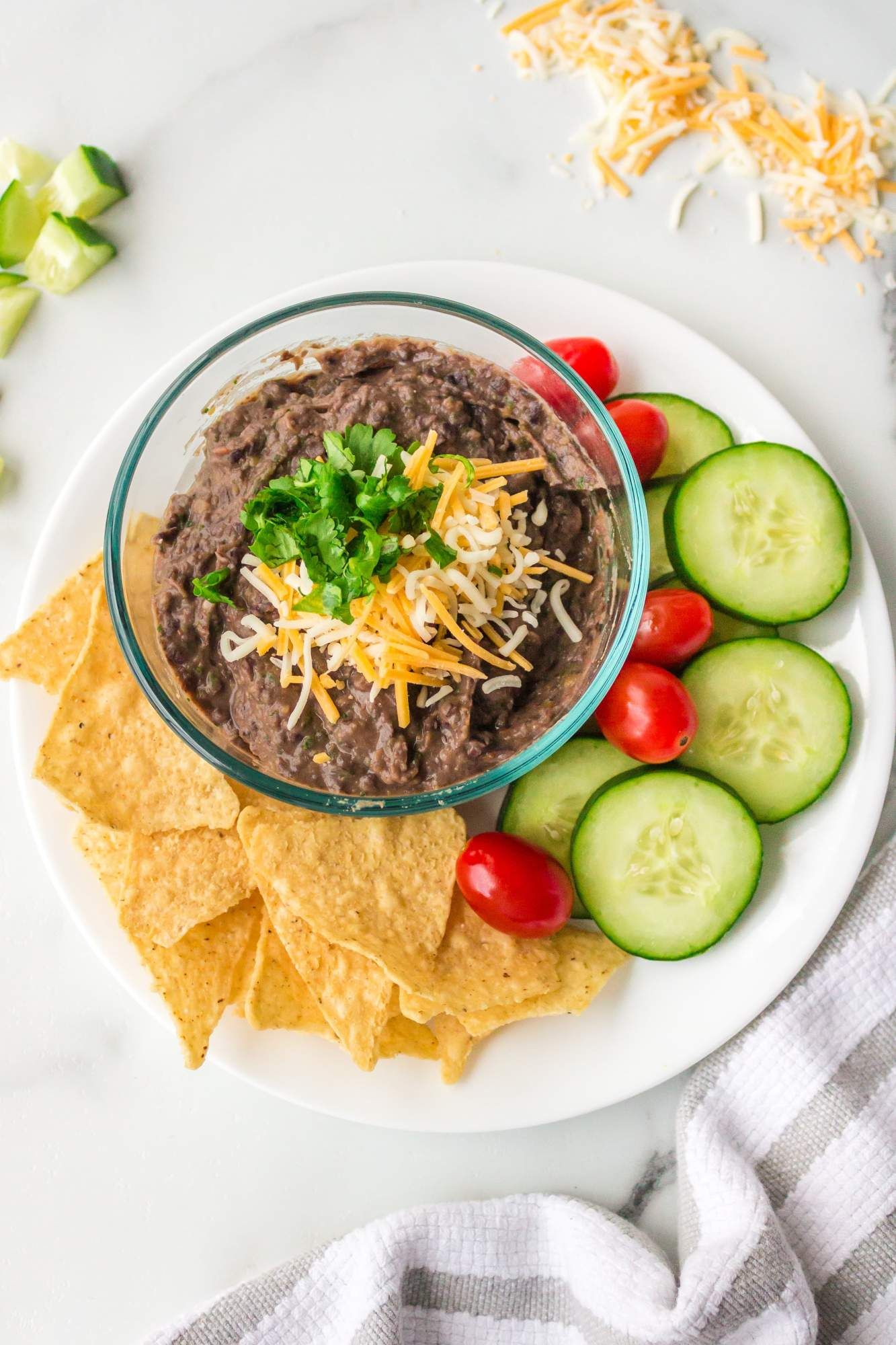 Bean dip made with black beans and salsa on a plate with cucumbers, tomatoes, baked tortilla chips, and cheese.