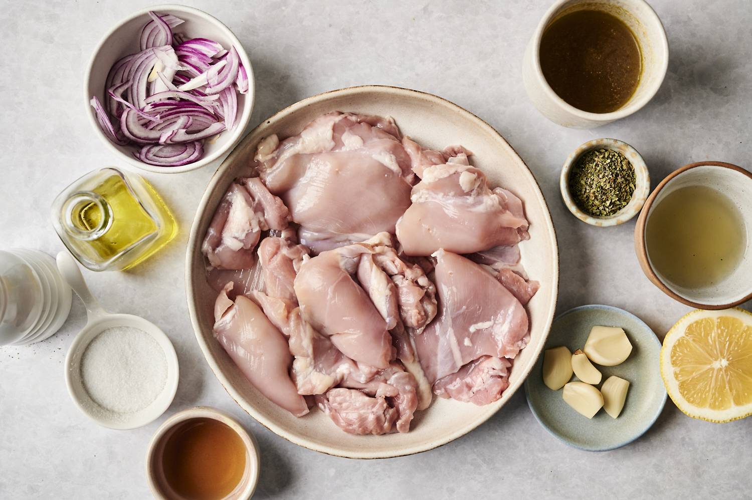 A flat lay of fresh ingredients for an easy Greek chicken dish, including raw chicken thighs, red onions, garlic, lemon, olive oil, seasonings, and broth, arranged on a light surface.