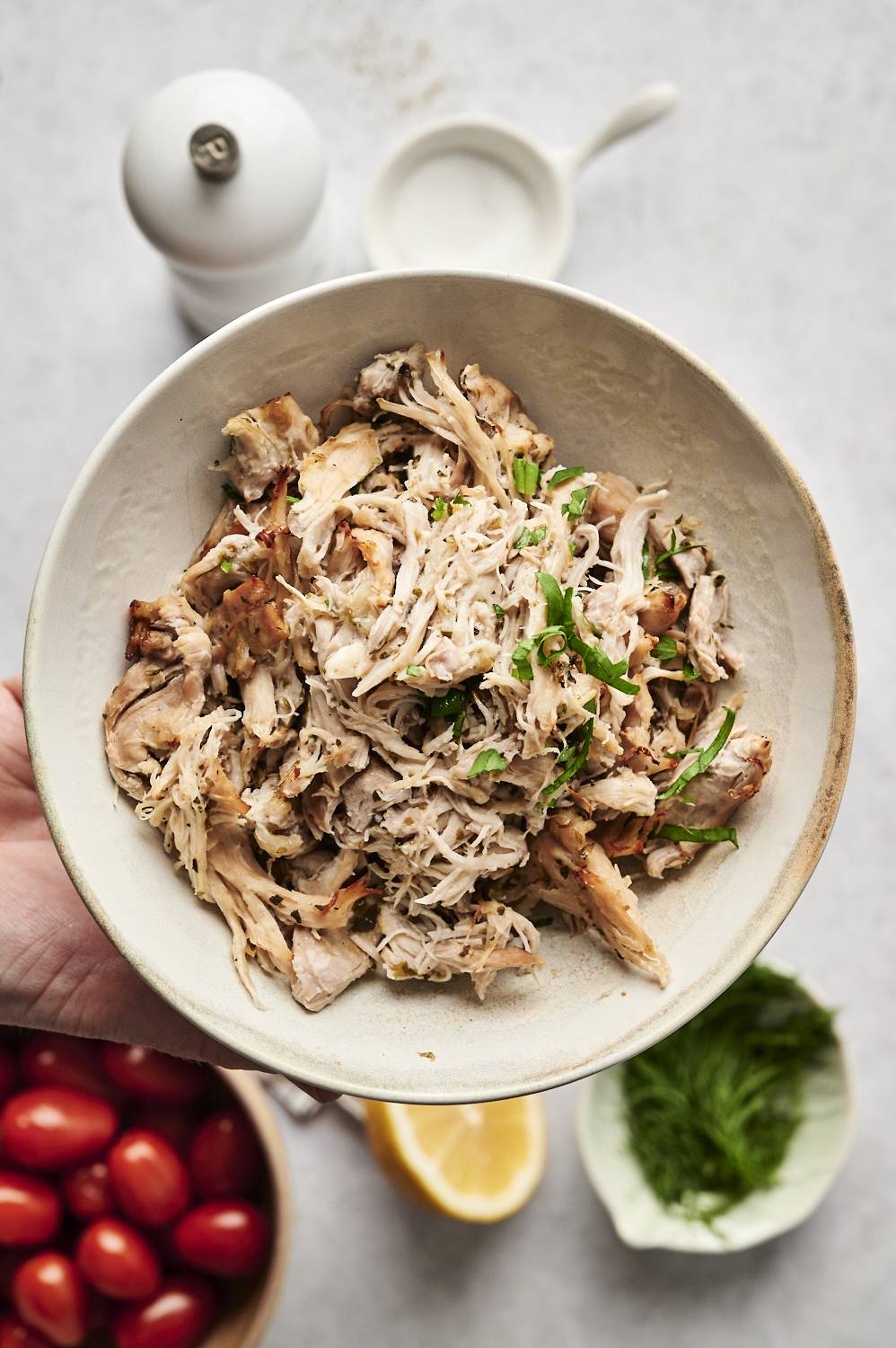A bowl of shredded Greek chicken, garnished with fresh herbs. The tender, juicy chicken has been slow-cooked or pressure-cooked with Mediterranean flavors. In the background, there are cherry tomatoes, fresh dill, lemon, and a salt dish, suggesting ingredients that pair well with the dish.