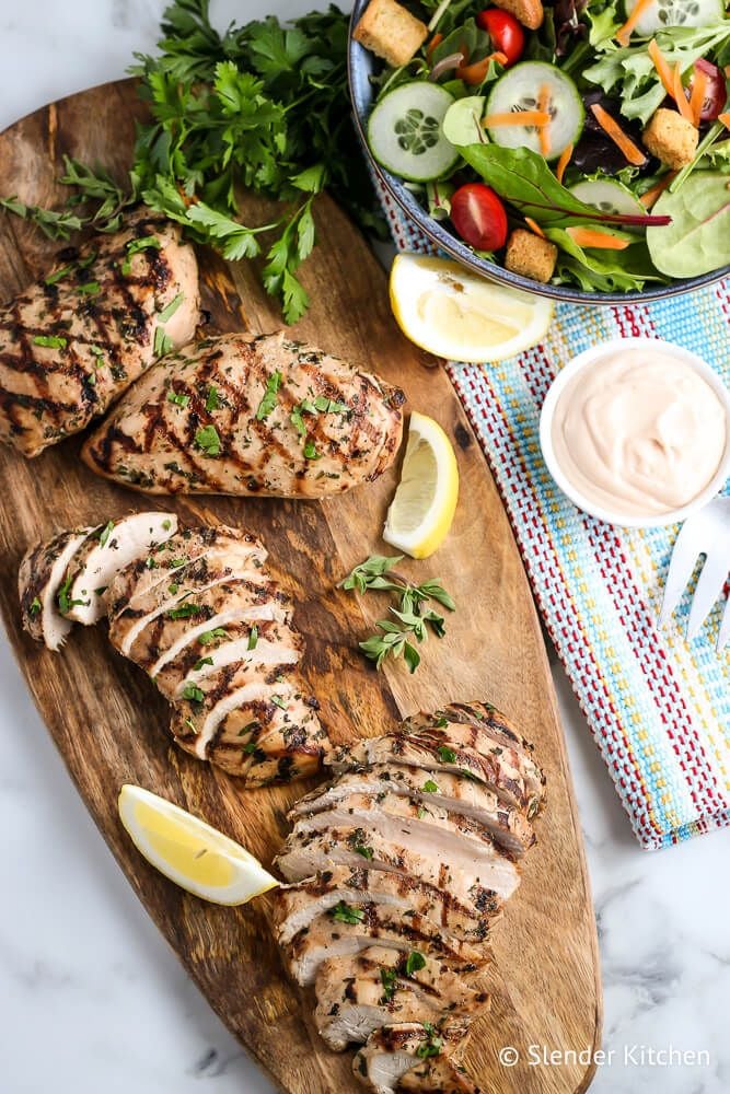 Chicken breasts that have been grilled sliced on a cutting board with salad and herbs on the side.