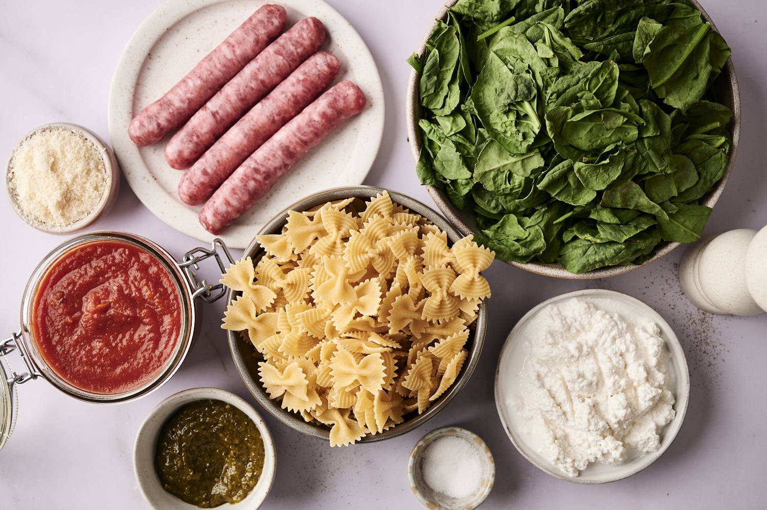 The ingredients of the high-protein pasta are arranged on a light-colored surface, including original Italian sausage, fresh spinach, bow and arrow pasta, ricotta cheese, marinara sauce, pesto, pesto, grated parmesan and a small bowl of salt.