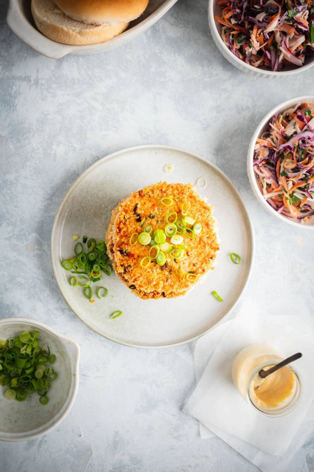 The salmon burger patty was breaded and served on a plate with coleslaw and burger buns on the side.