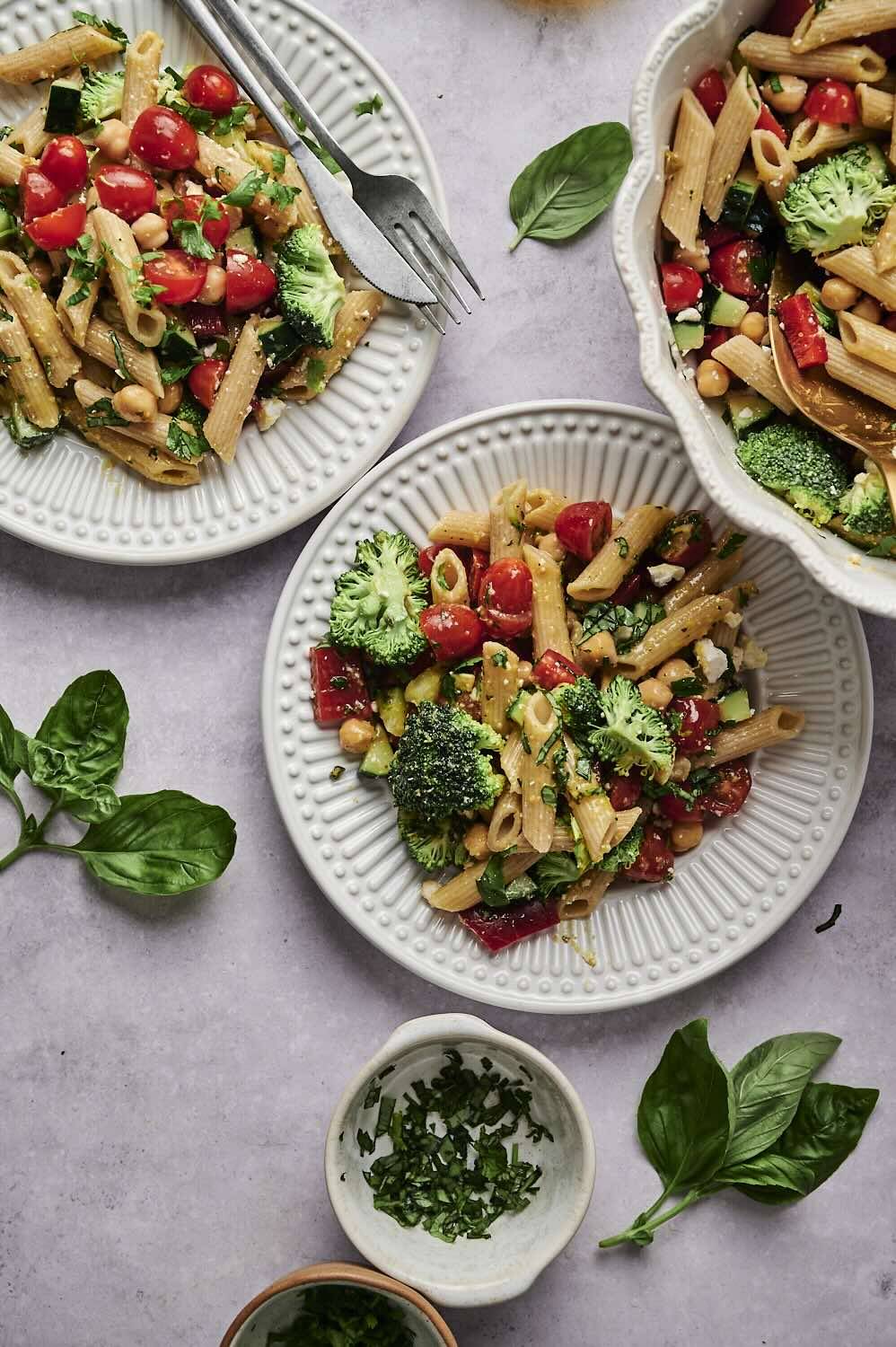 A vibrant bowl of pasta salad with cherry tomatoes, broccoli, and chickpeas, garnished with fresh herbs on textured plates.