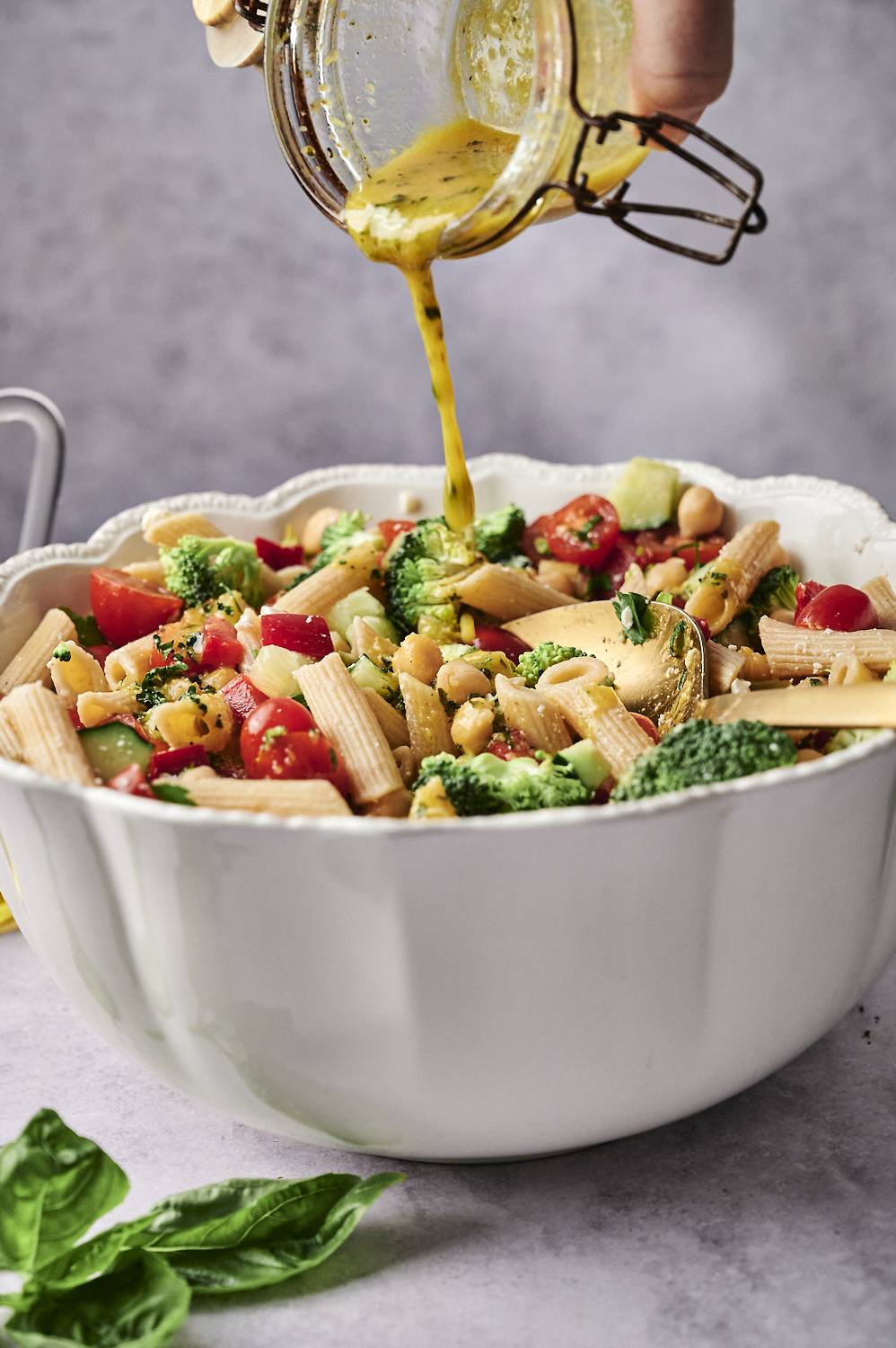 A hand pours a vibrant yellow dressing over a colorful salad of pasta, cherry tomatoes, broccoli, and chickpeas in a large white bowl.