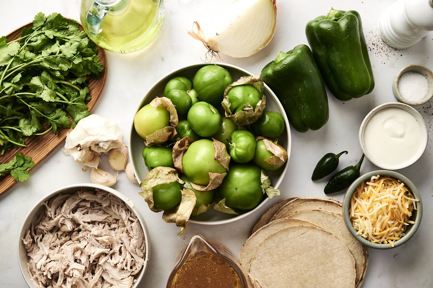 A variety of ingredients for enchiladas suizas laid out, including cilantro, green bell peppers, tomatillos, garlic, shredded chicken, tortillas, shredded cheese, and sour cream.