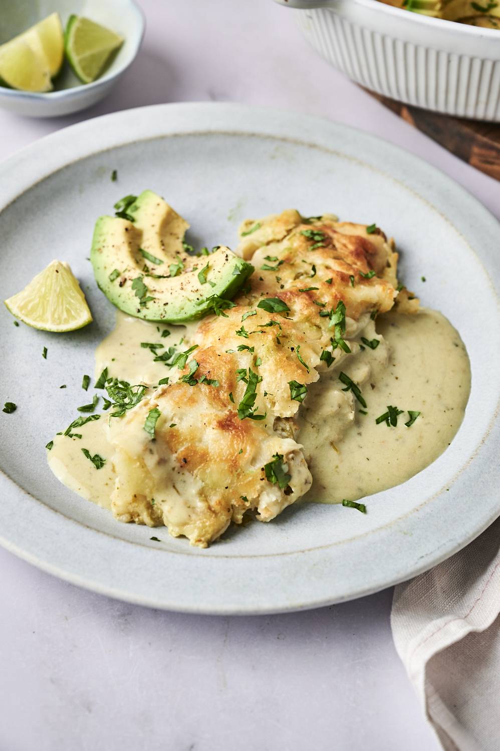 A plate of creamy chicken enchiladas topped with cheese and cilantro, served with avocado slices and lime wedges.