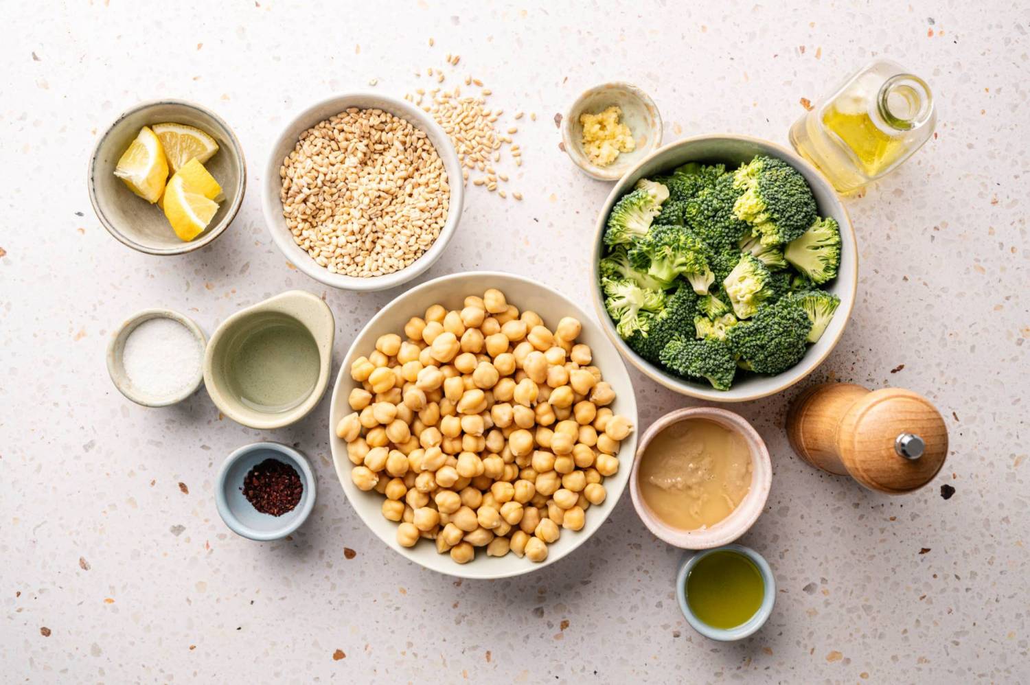 Assorted ingredients on speckled countertop include chickpeas, broccoli, sesame seeds, lemon wedges, olive oil, garlic and tahini