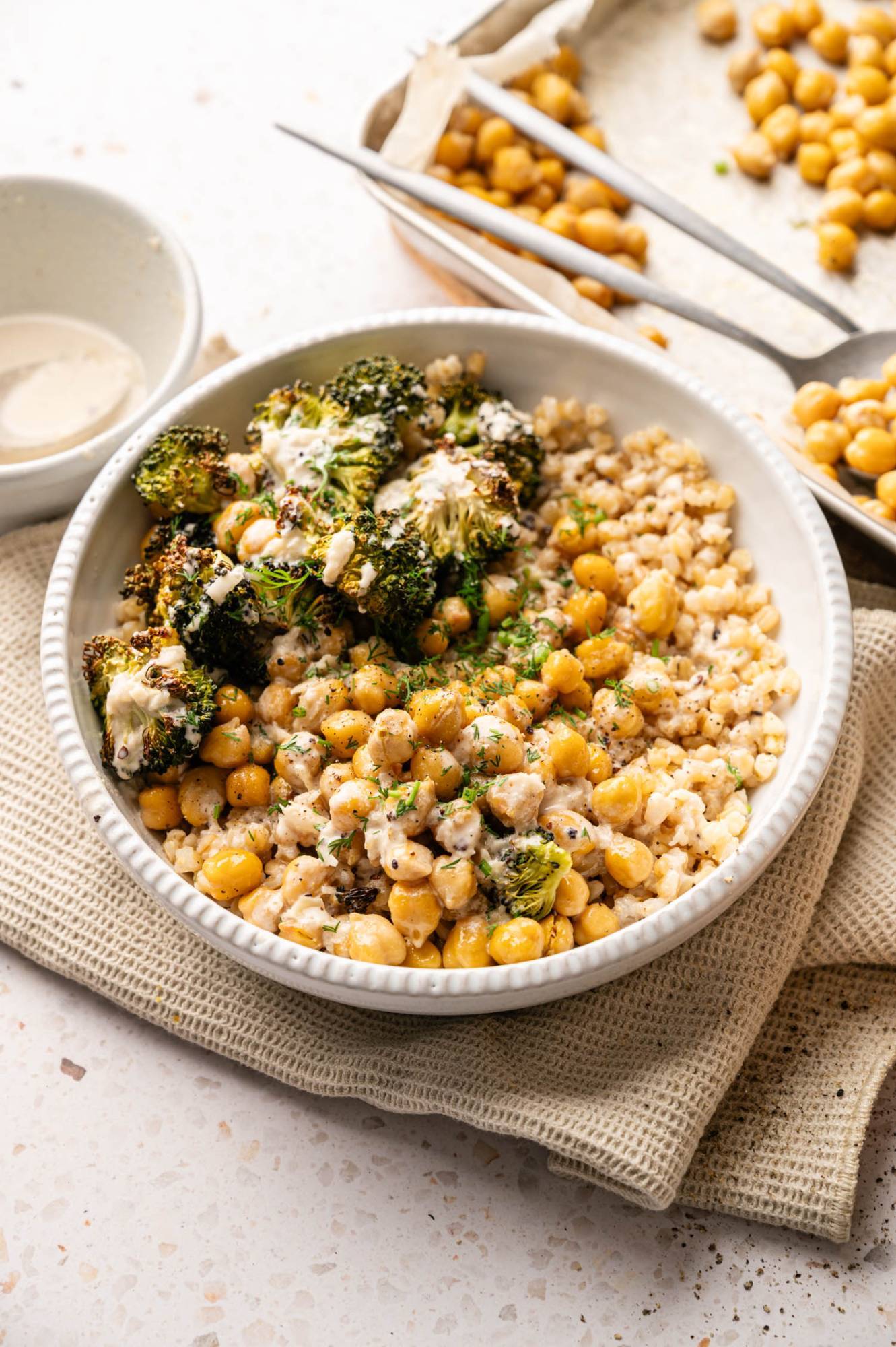 A bowl of roasted chickpeas, broccoli and grains topped with a creamy sauce. Next to it is a small plate of sauce and a tray with more chickpeas.