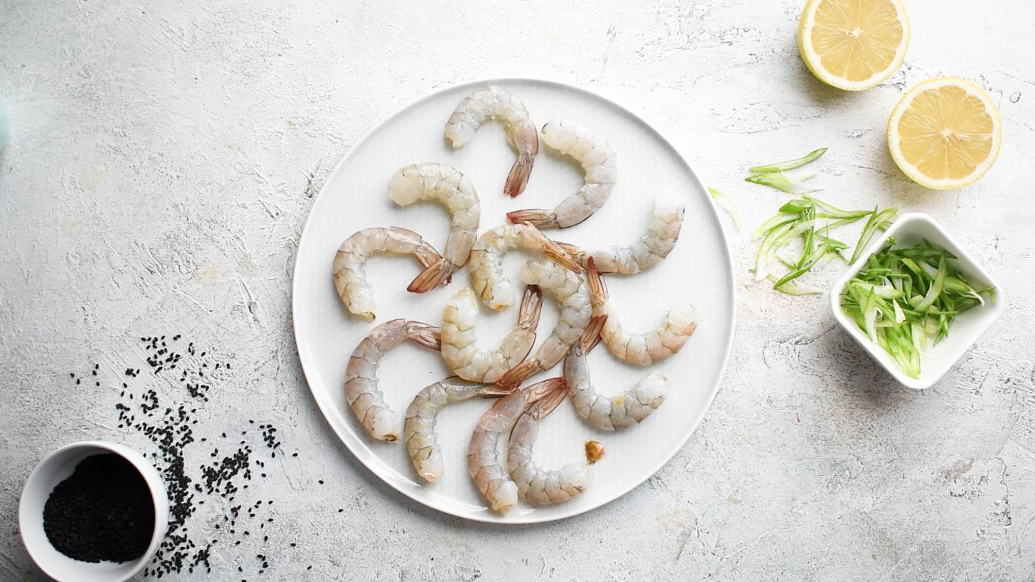 A plate of raw, peeled shrimp sits on a white countertop with bowls of black sesame seeds, lemons, and sliced scallions.