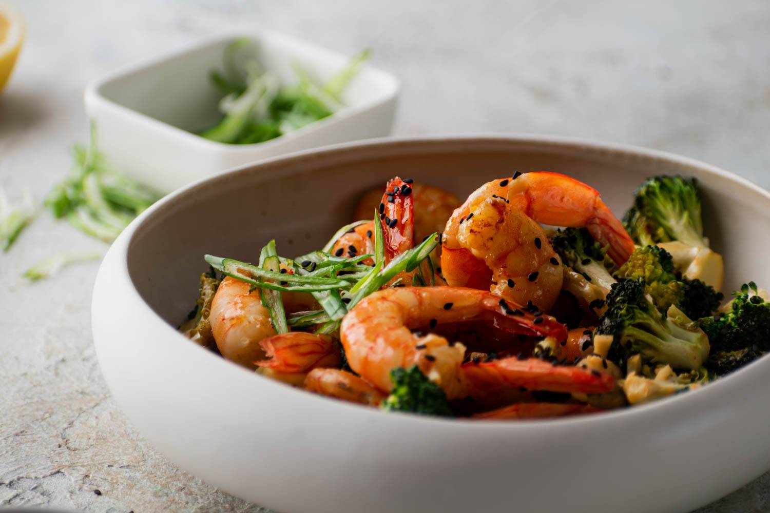 A bowl of firecracker shrimp and broccoli, garnished with sliced scallions and sesame seeds.