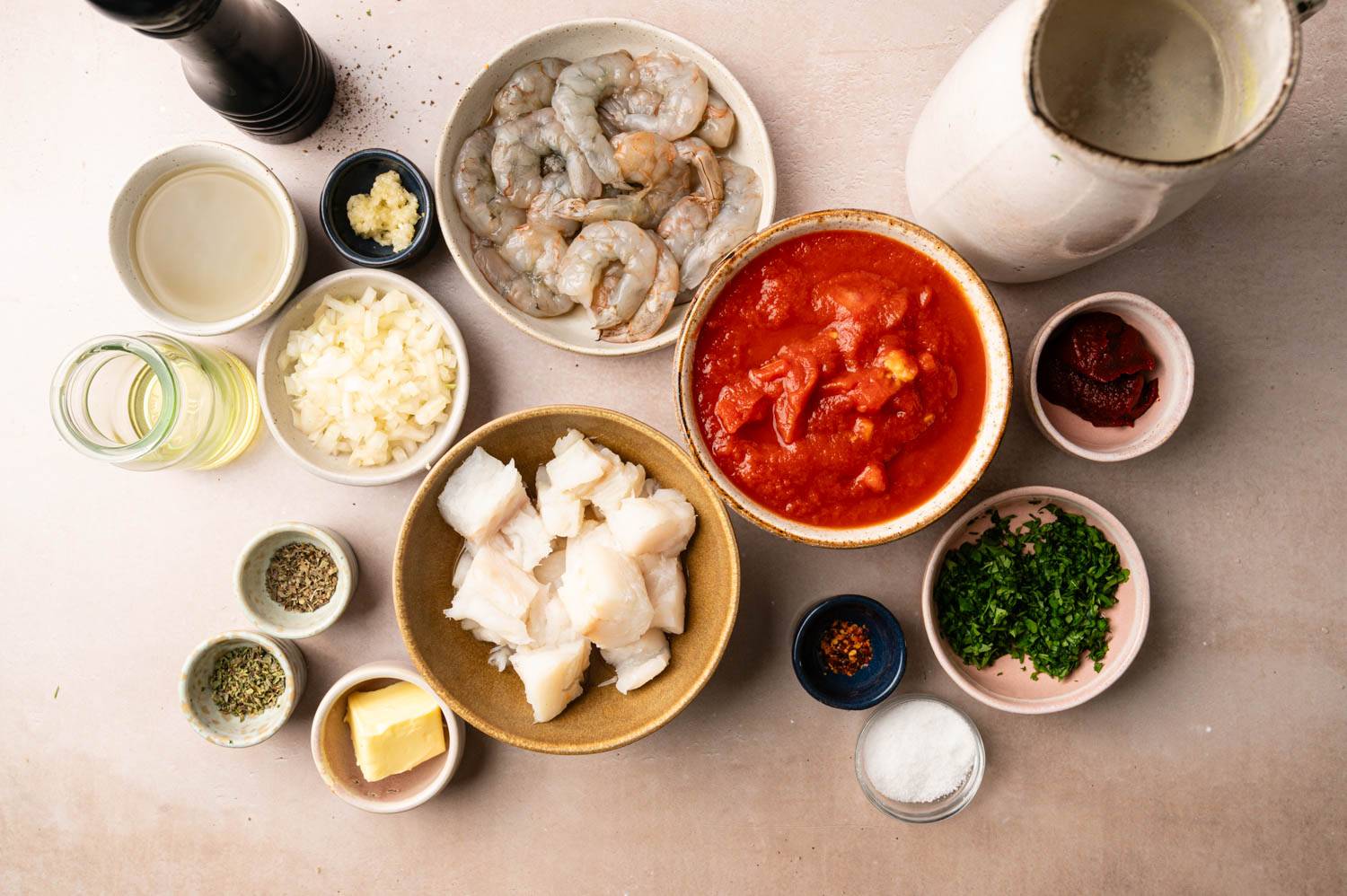 Ingredients for seafood dish on a table: raw shrimp, cubed fish, tomato sauce, chopped herbs, onions, garlic, spices, butter, and broth. Cooking preparation.