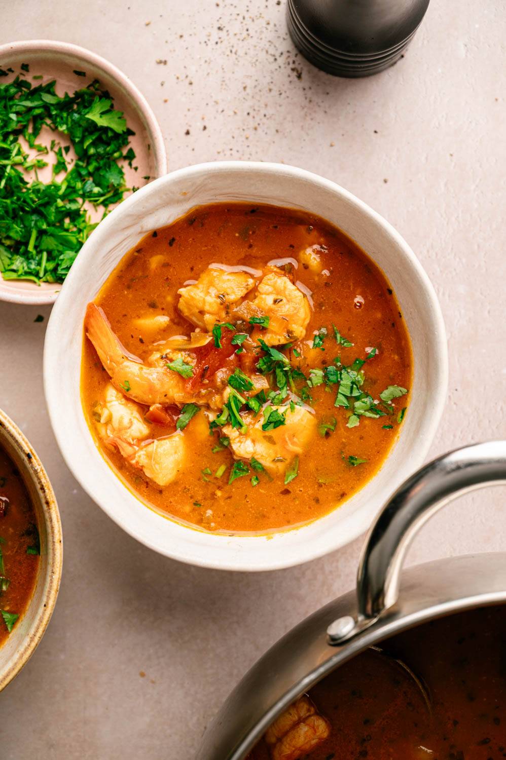 A bowl of shrimp bisque garnished with fresh parsley. Rich, orange broth with shrimp pieces, surrounded by a small dish of parsley and a pepper grinder.