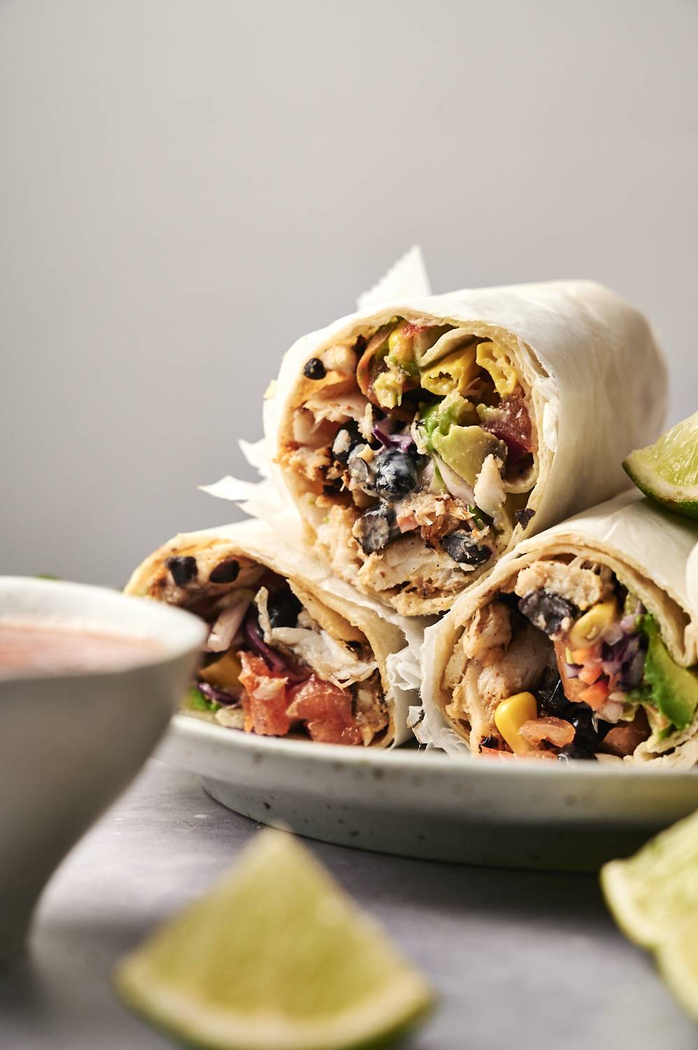 Three delicious burritos, filled with chicken, black beans, and fresh vegetables, served with a lime wedge and dipping sauce.