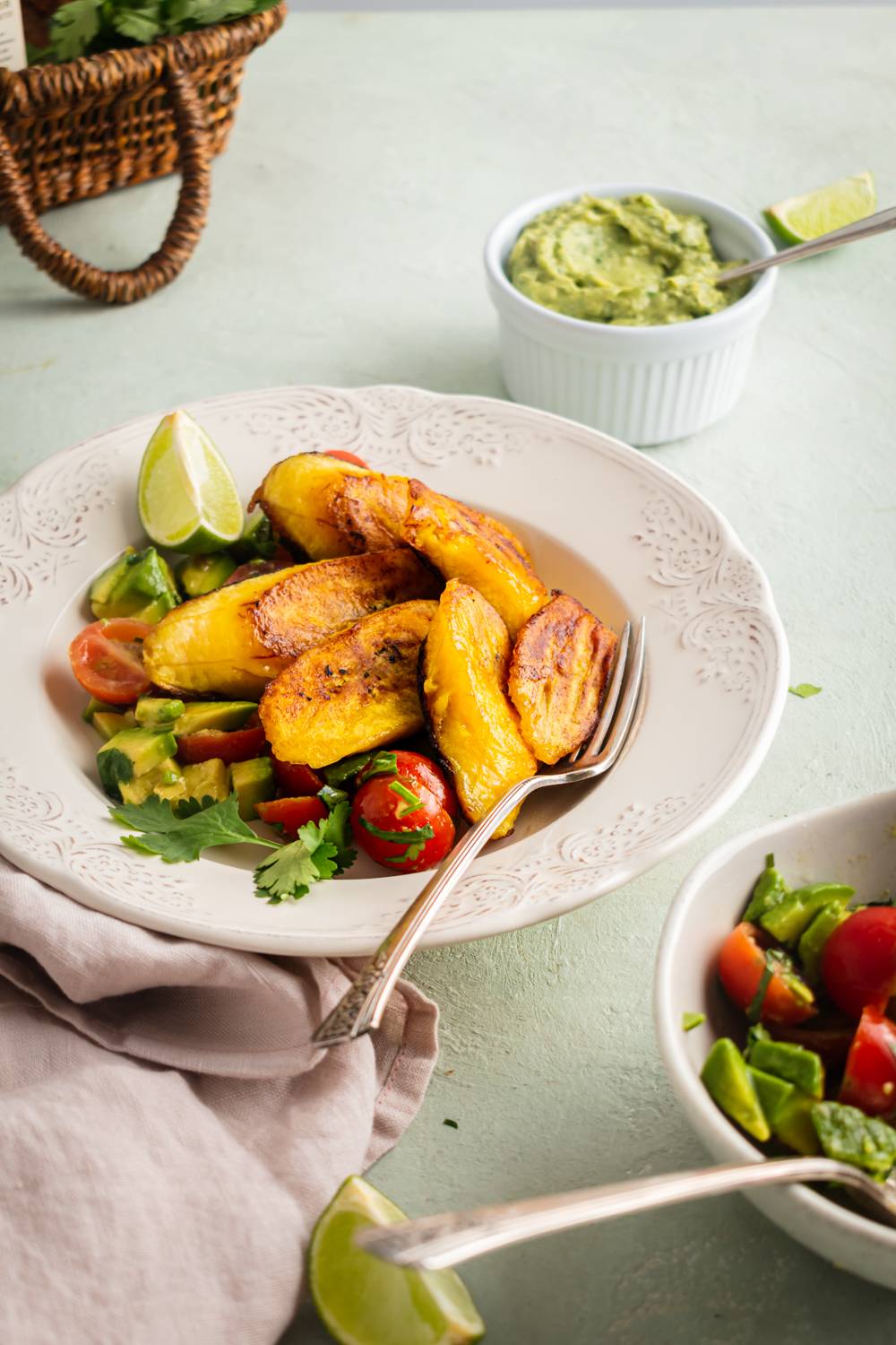 A plate of golden fried plantains with lime on top of a cherry tomato and avocado salad, served with a bowl of guacamole.