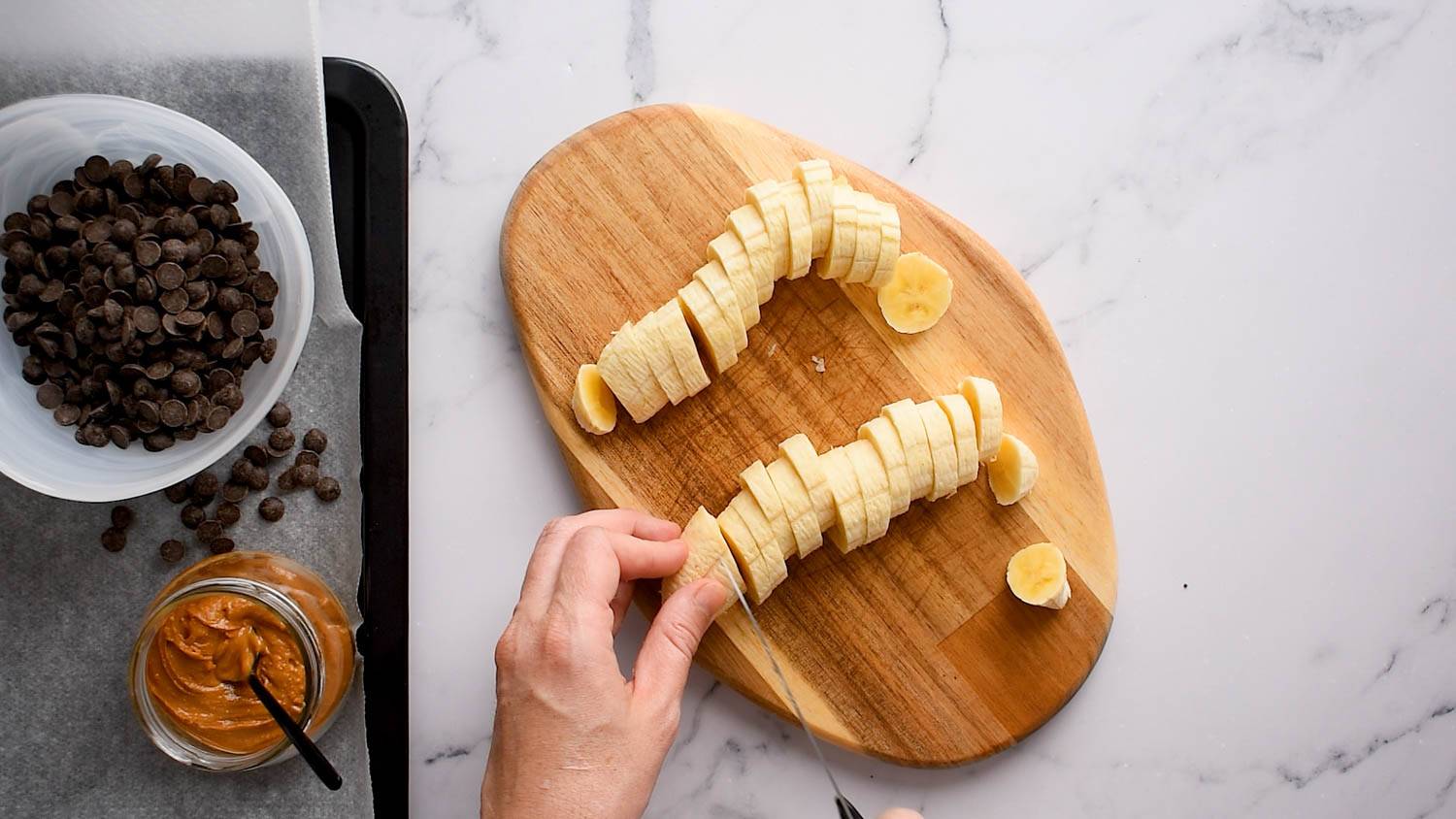 A hand slices a bunch of banana pieces on a wooden cutting board, surrounded by chocolate chips and peanut butter.