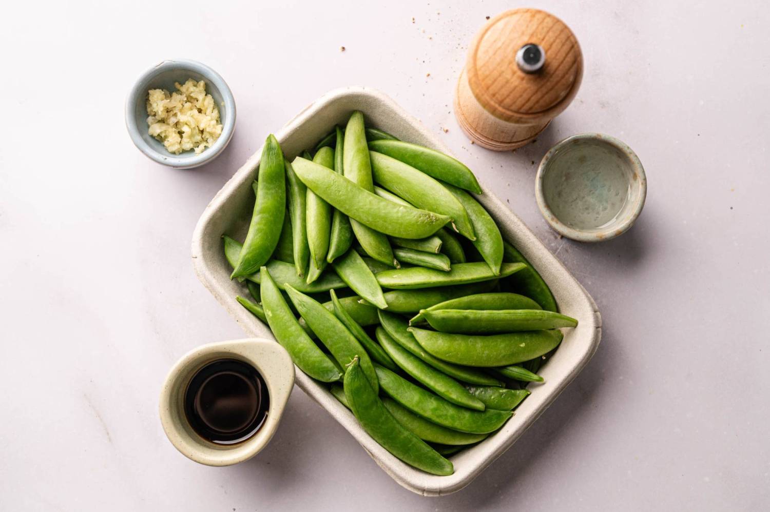 Fresh green sugar snap peas in a white dish, surrounded by garlic, soy sauce, and a wooden pepper grinder on a light surface.