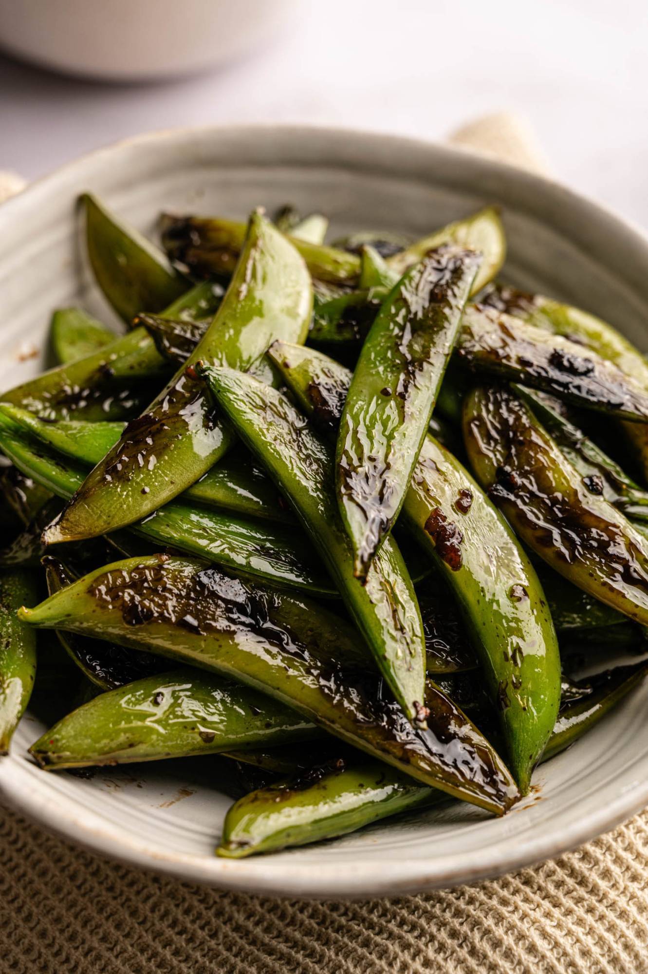 A white bowl filled with vibrant green sugar snap peas glazed with a shiny, savory sauce.