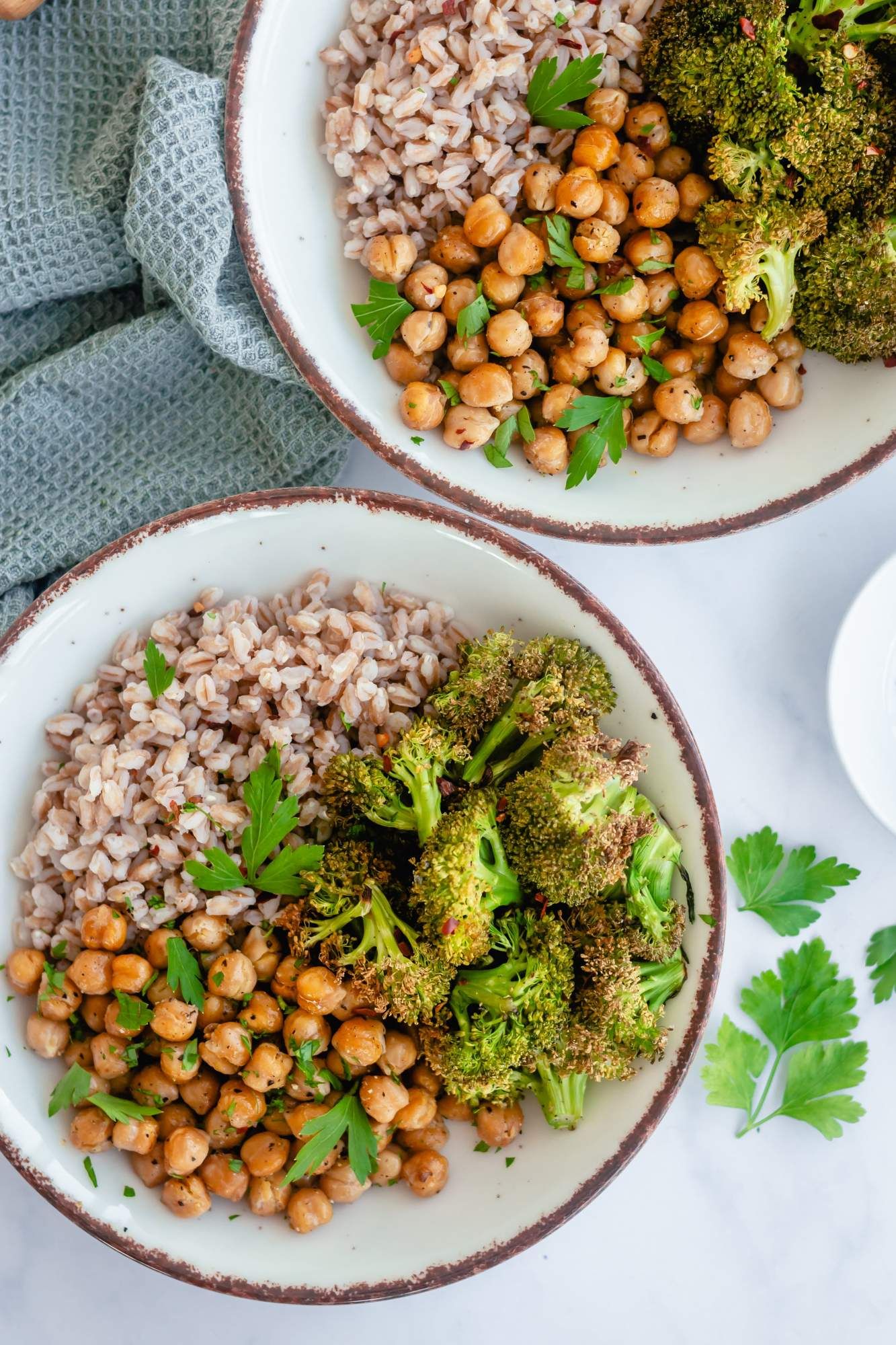 Chickpea and broccoli farro bowls with farro and garlic tahini sauce in two bowls with a blue napkin.