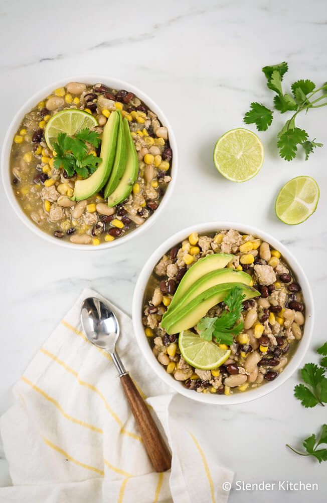 Green chili with ground turkey in two bowls with corn, beans, ground turkey, avocado, lime, and cilantro.