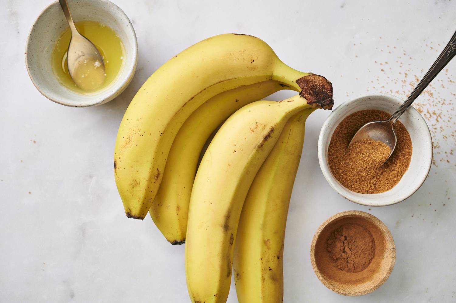 A bunch of ripe bananas alongside bowls of honey, brown sugar, and ground cinnamon on a marble countertop.