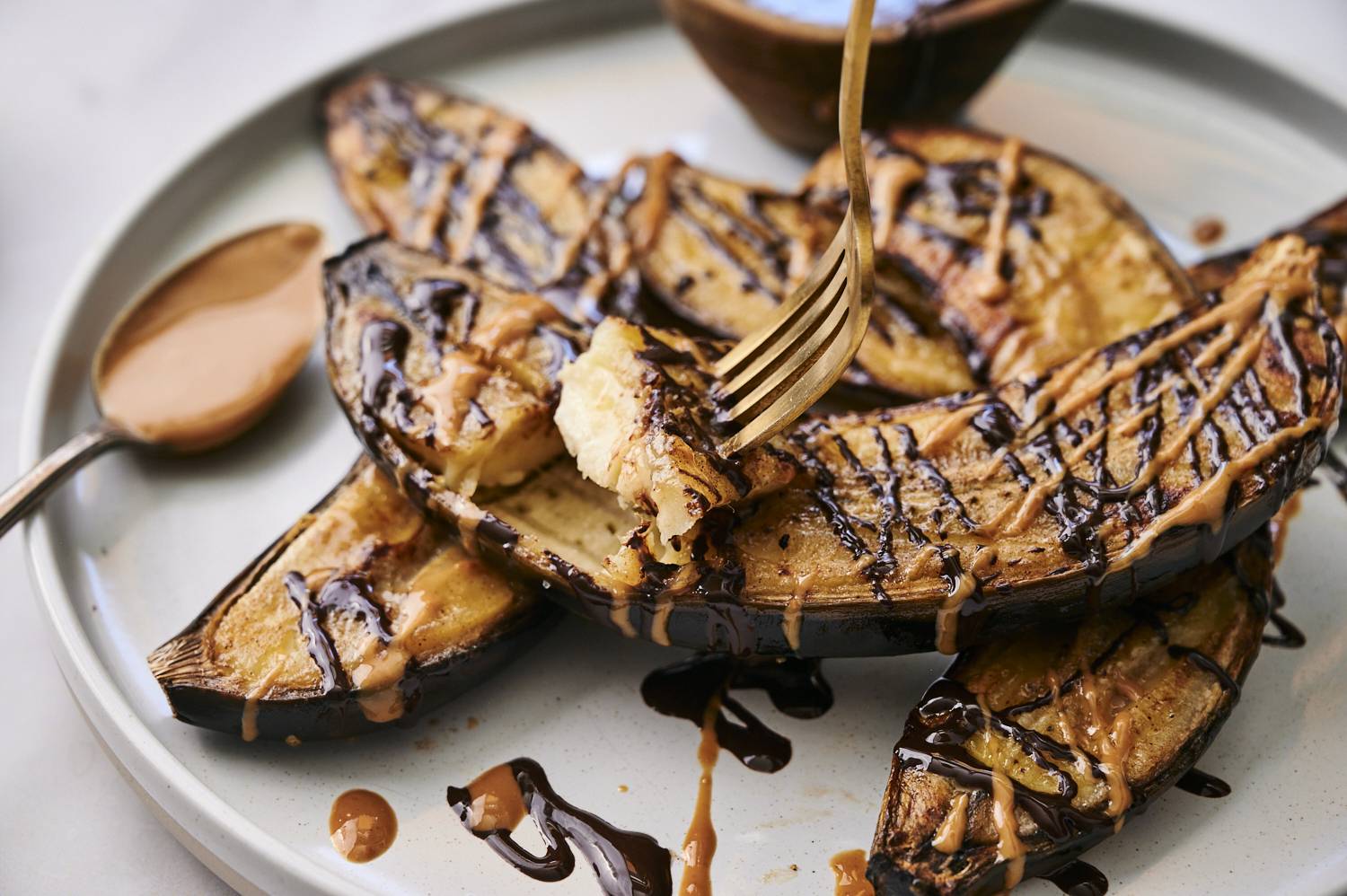 Grilled bananas drizzled with chocolate and peanut butter, served on a round plate with a spoon nearby.