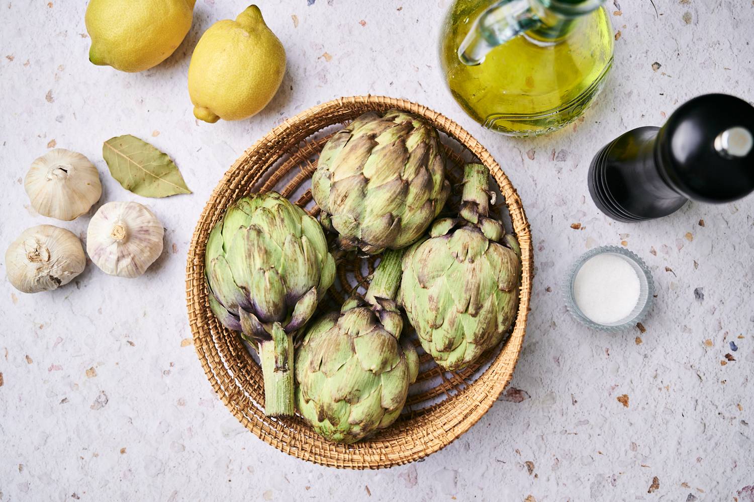 Ingredients for grilled artichokes including raw artichokes, lemon, garlic, olive oil, salt, and pepper.