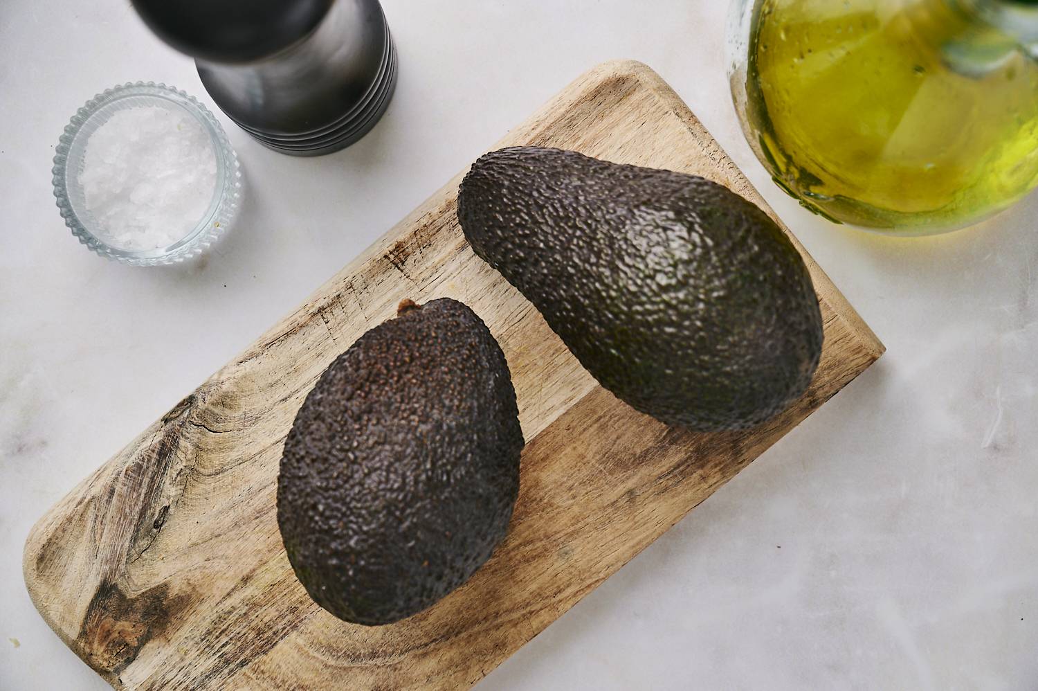 Two ripe avocados on a wooden cutting board, alongside a glass bowl of salt and a bottle of olive oil.