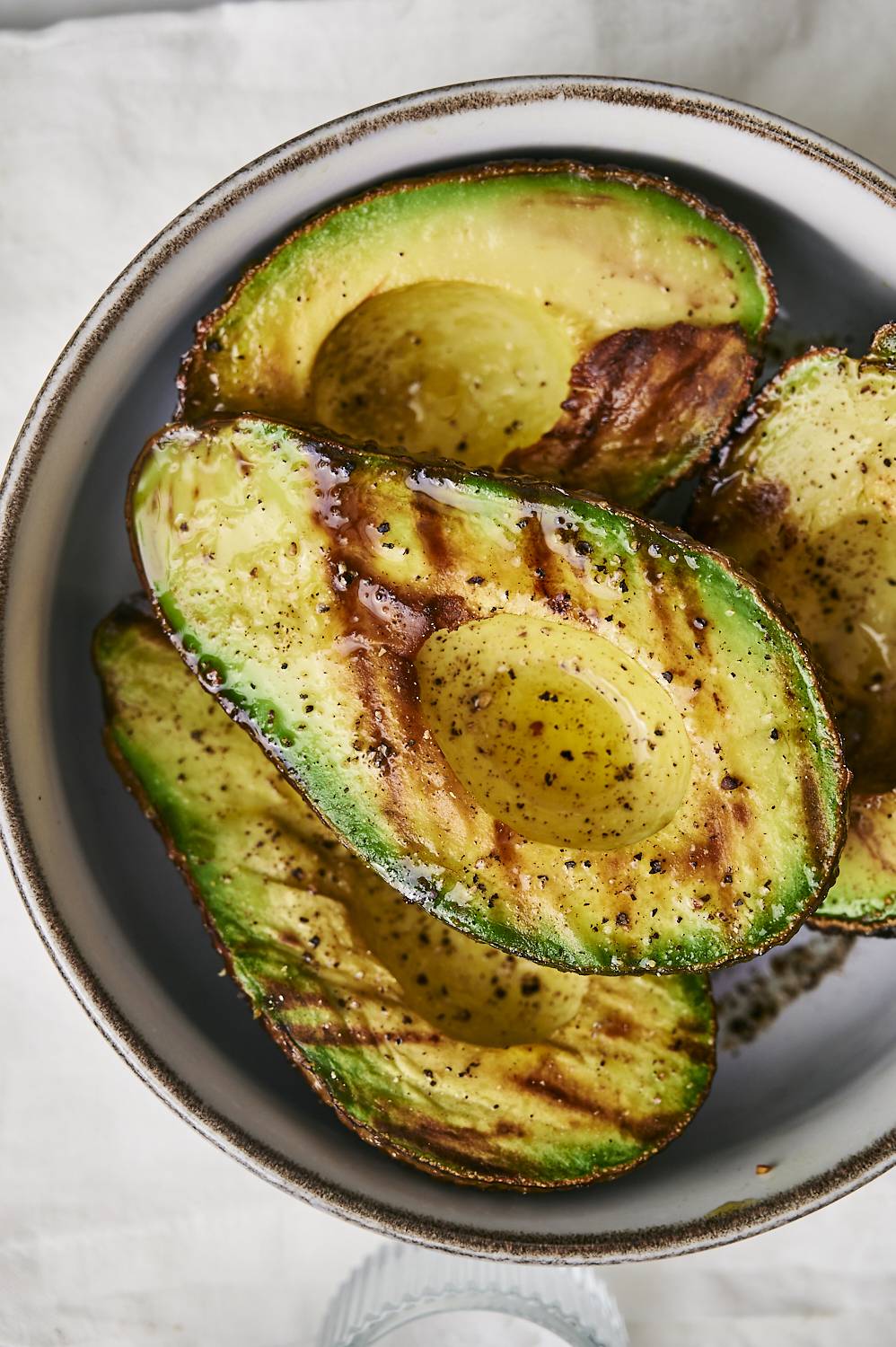 Grilled avocado halves drizzled with olive oil and sprinkled with black pepper in a gray bowl, resting on a light linen background.