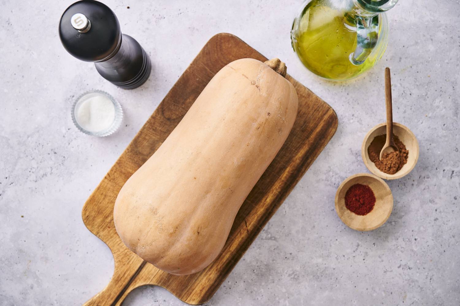 A whole butternut squash on a wooden cutting board, surrounded by spices, salt, and a bottle of olive oil.
