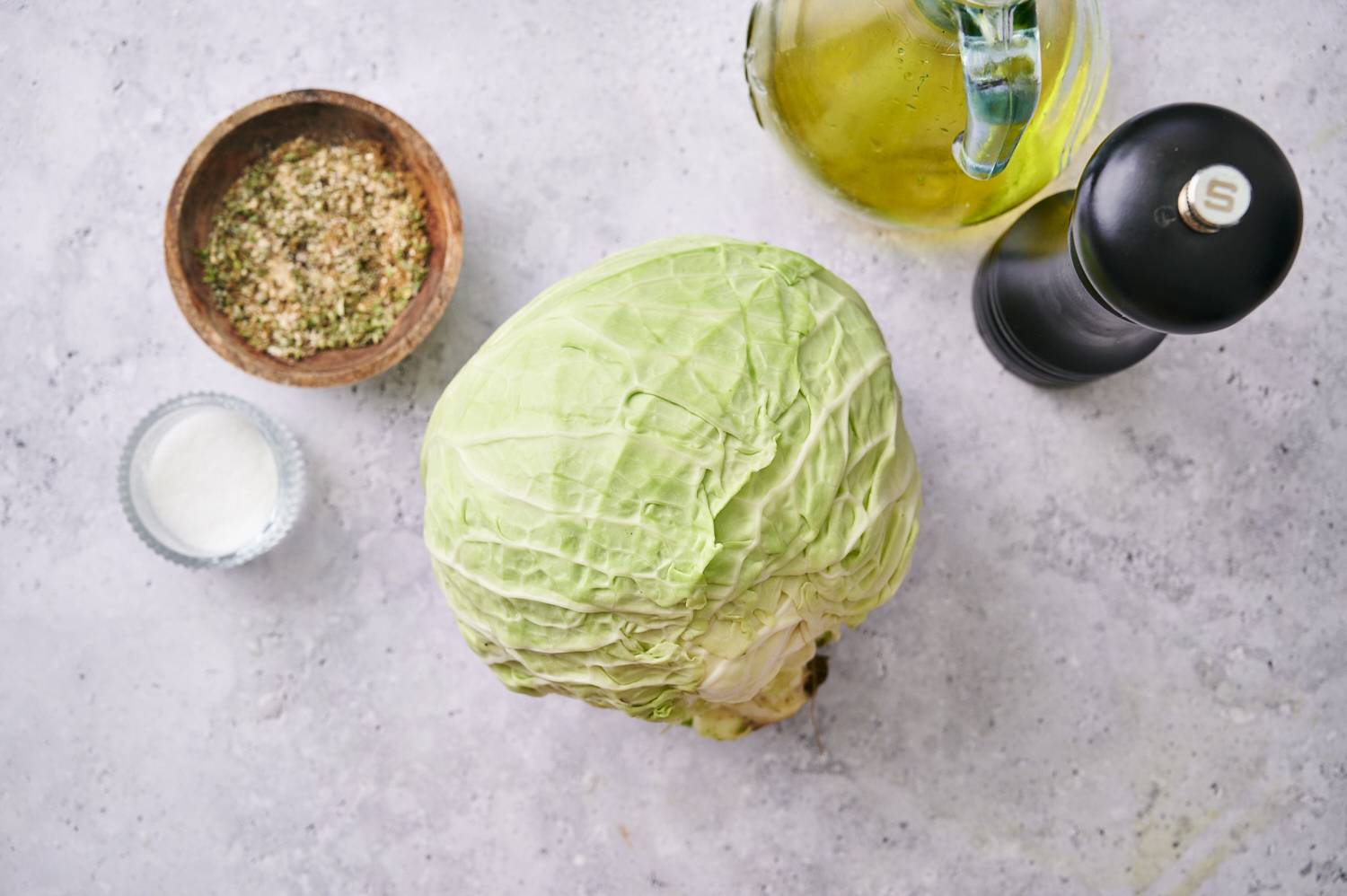 A fresh green cabbage sits on a light surface beside a bowl of seasoning, salt, olive oil, and a pepper grinder.