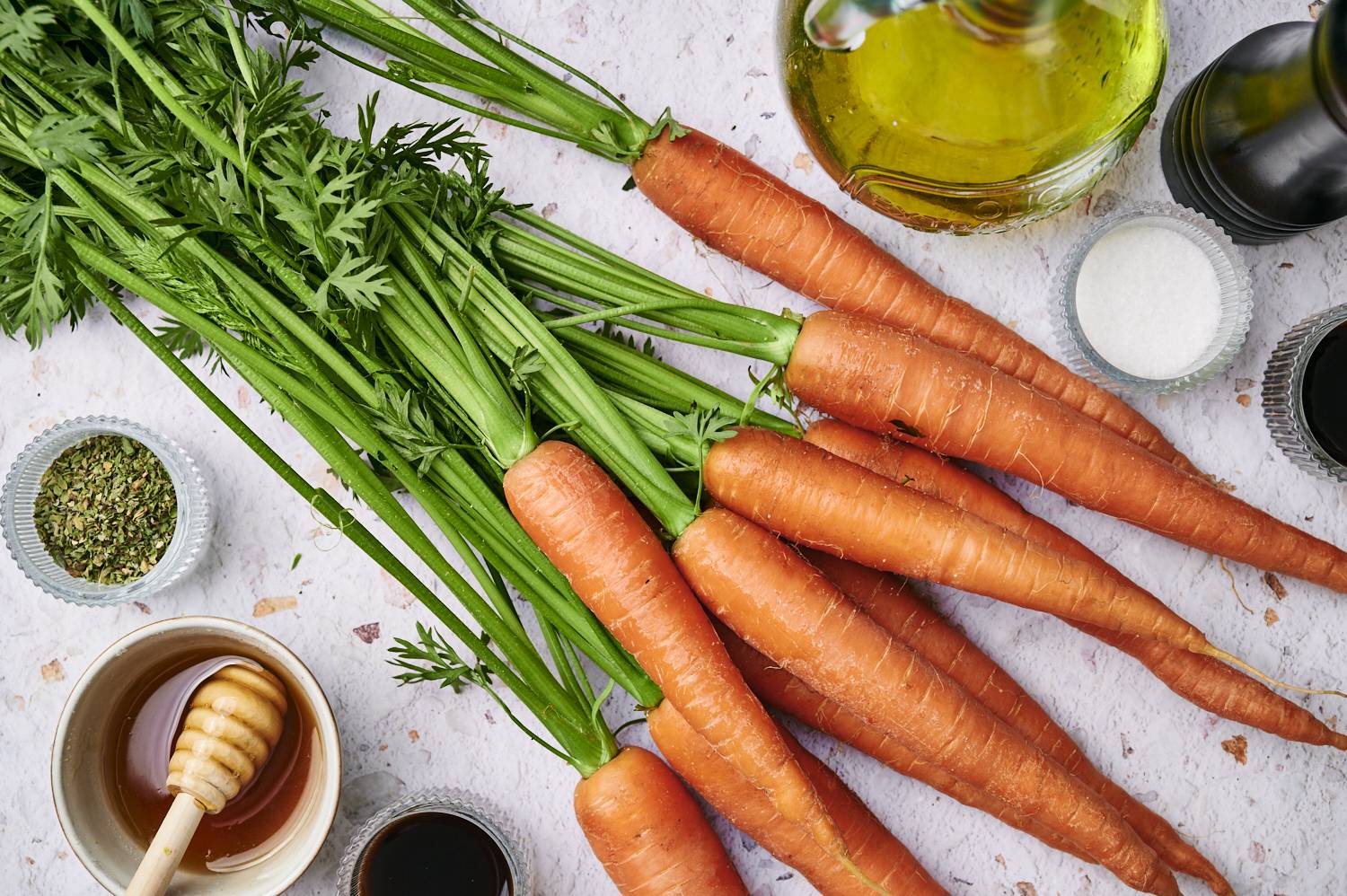 A rustic arrangement of fresh carrots with green tops, surrounded by olive oil, honey, herbs, salt, and balsamic vinegar.