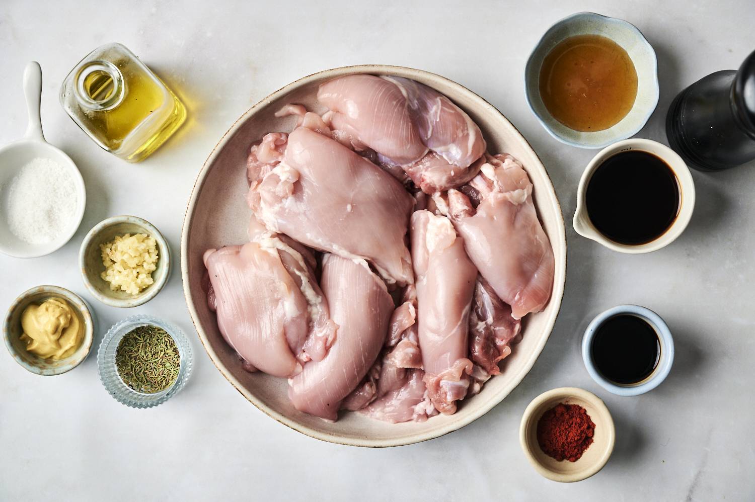 A bowl of raw chicken thighs surrounded by bowls of ingredients: oil, vinegar, mustard, garlic, herbs, and spices on a light countertop.