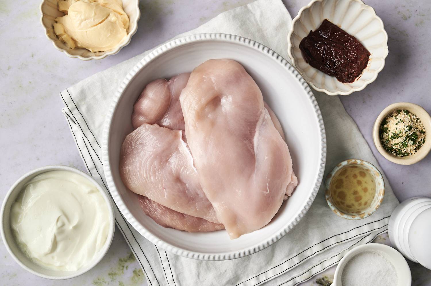 Raw chicken breasts in a bowl surrounded by ingredients like yogurt, mustard, chipotle sauce, spices, and oil on a textured cloth.