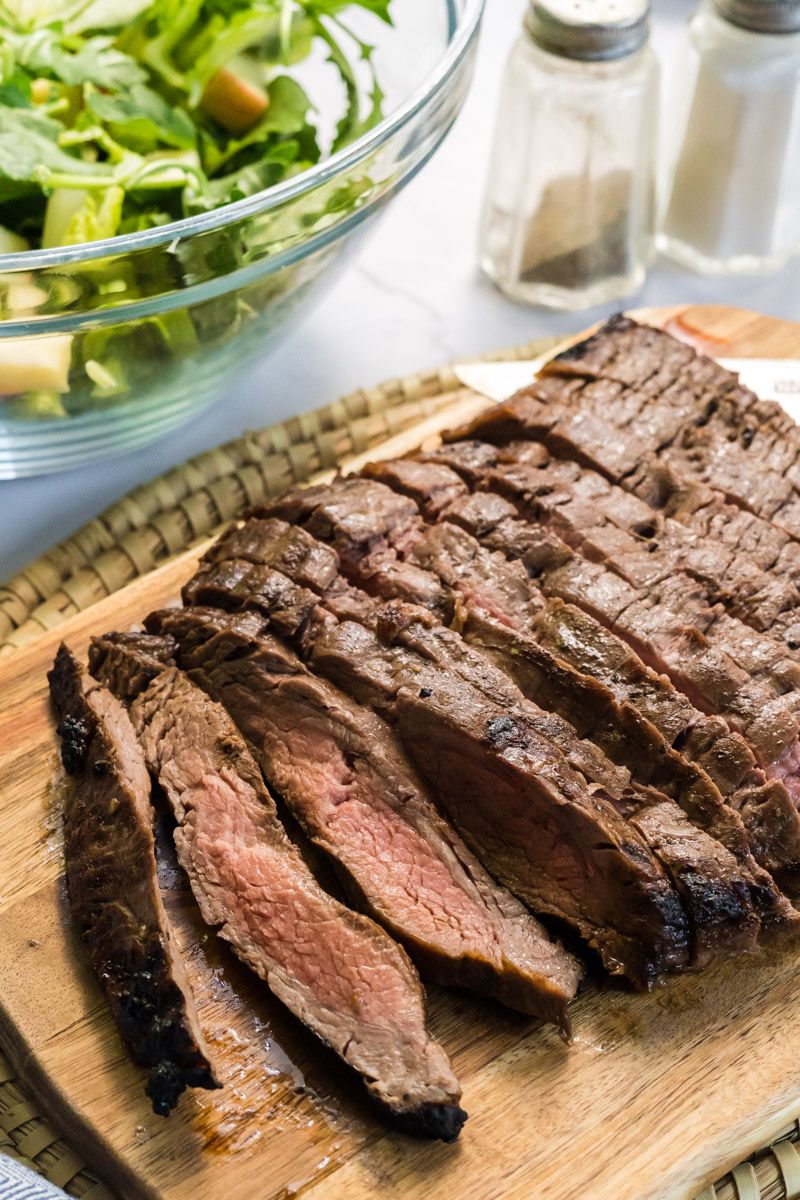 Grilled balsamic garlic flank steak sliced on a cutting board with salad in the background.