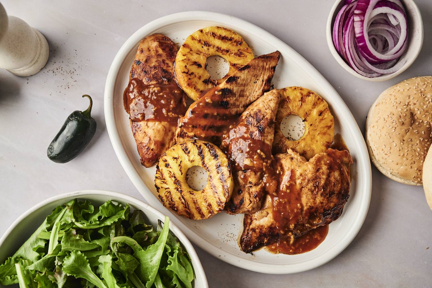 Grilled chicken breasts with BBQ sauce and grilled pineapple slices on a platter, alongside fresh salad leaves, red onion, and burger buns.