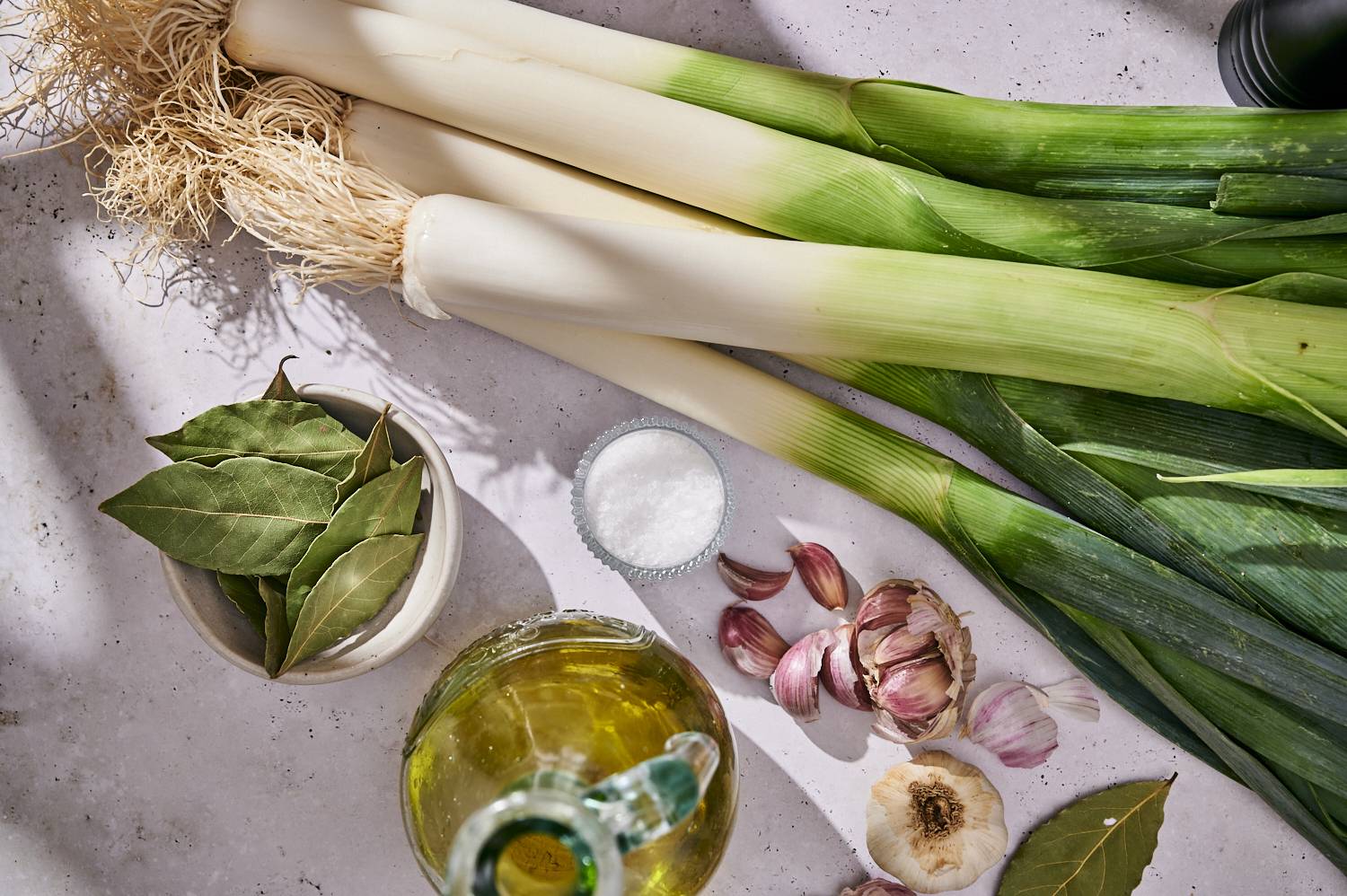 Fresh leeks with garlic, bay leaves, olive oil, and salt arranged on a light countertop, ready for grilling preparation.