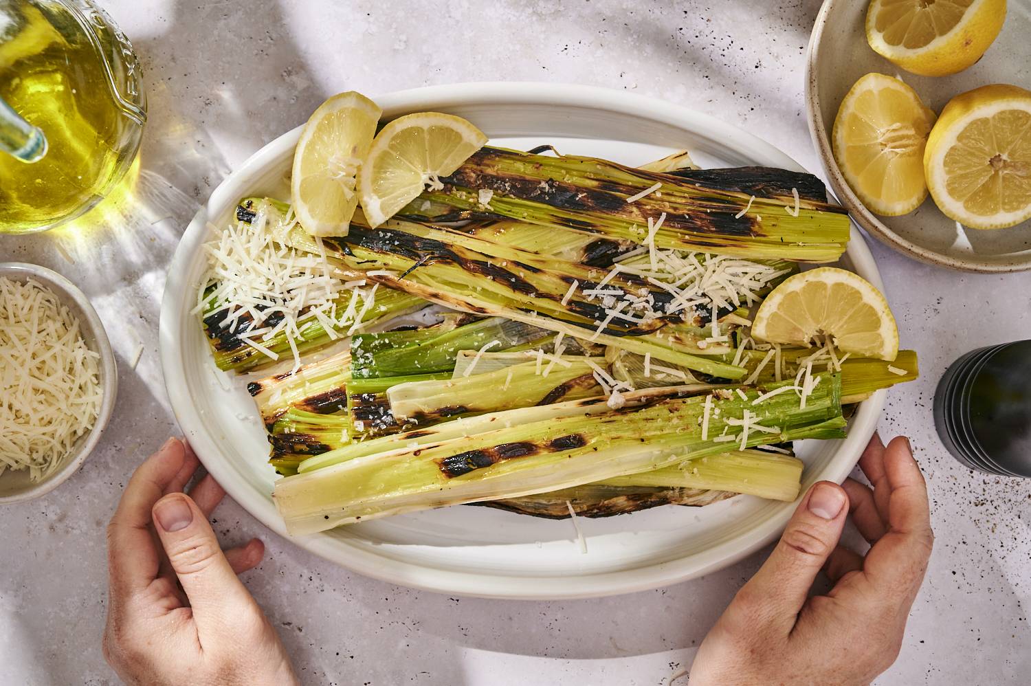 Grilled leeks with char marks, topped with grated Parmesan cheese and served with lemon wedges on a white plate, with a drizzle of olive oil nearby.