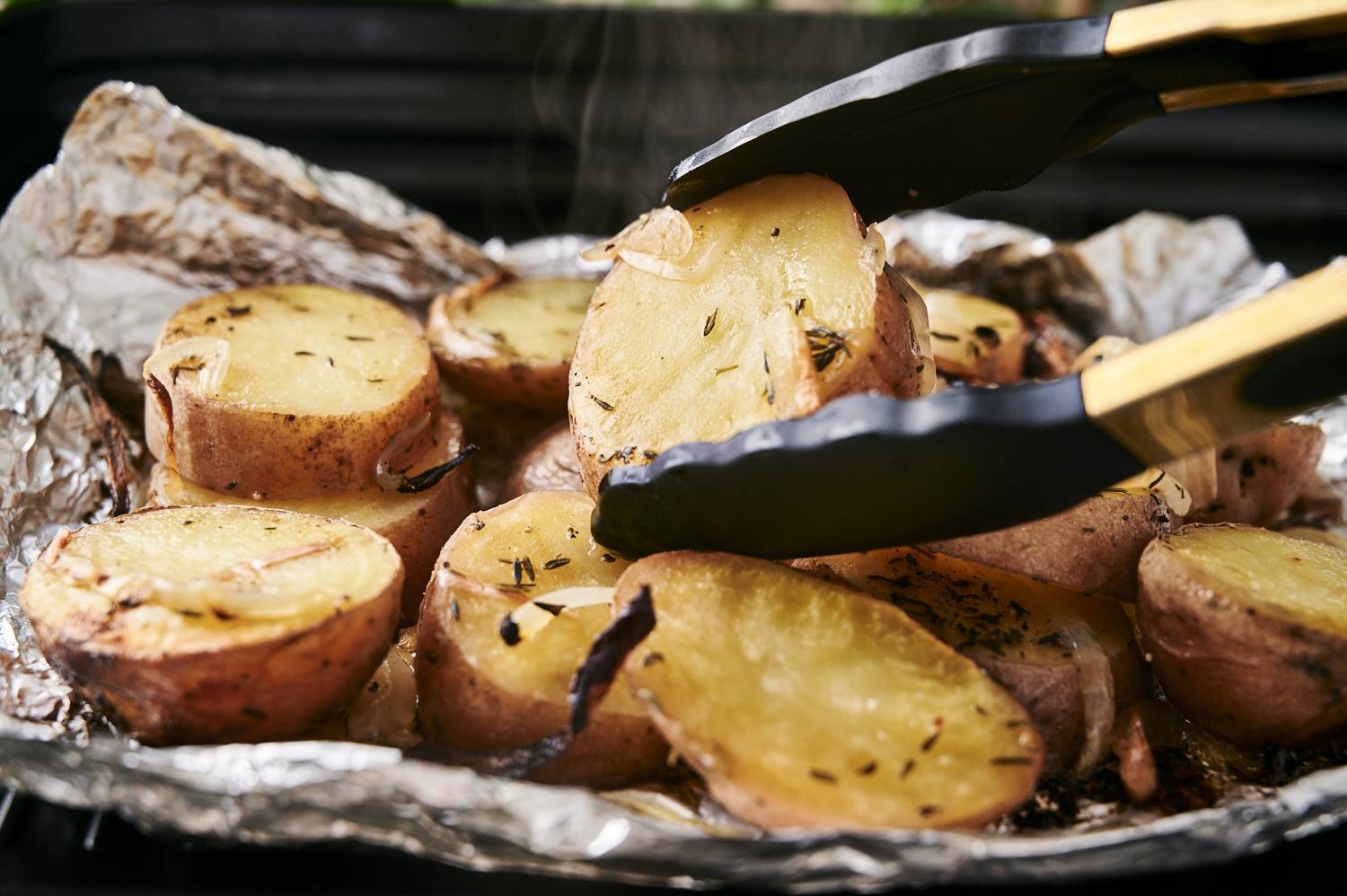 Tongs picking up a grilled red potato from a foil packet on the grill.