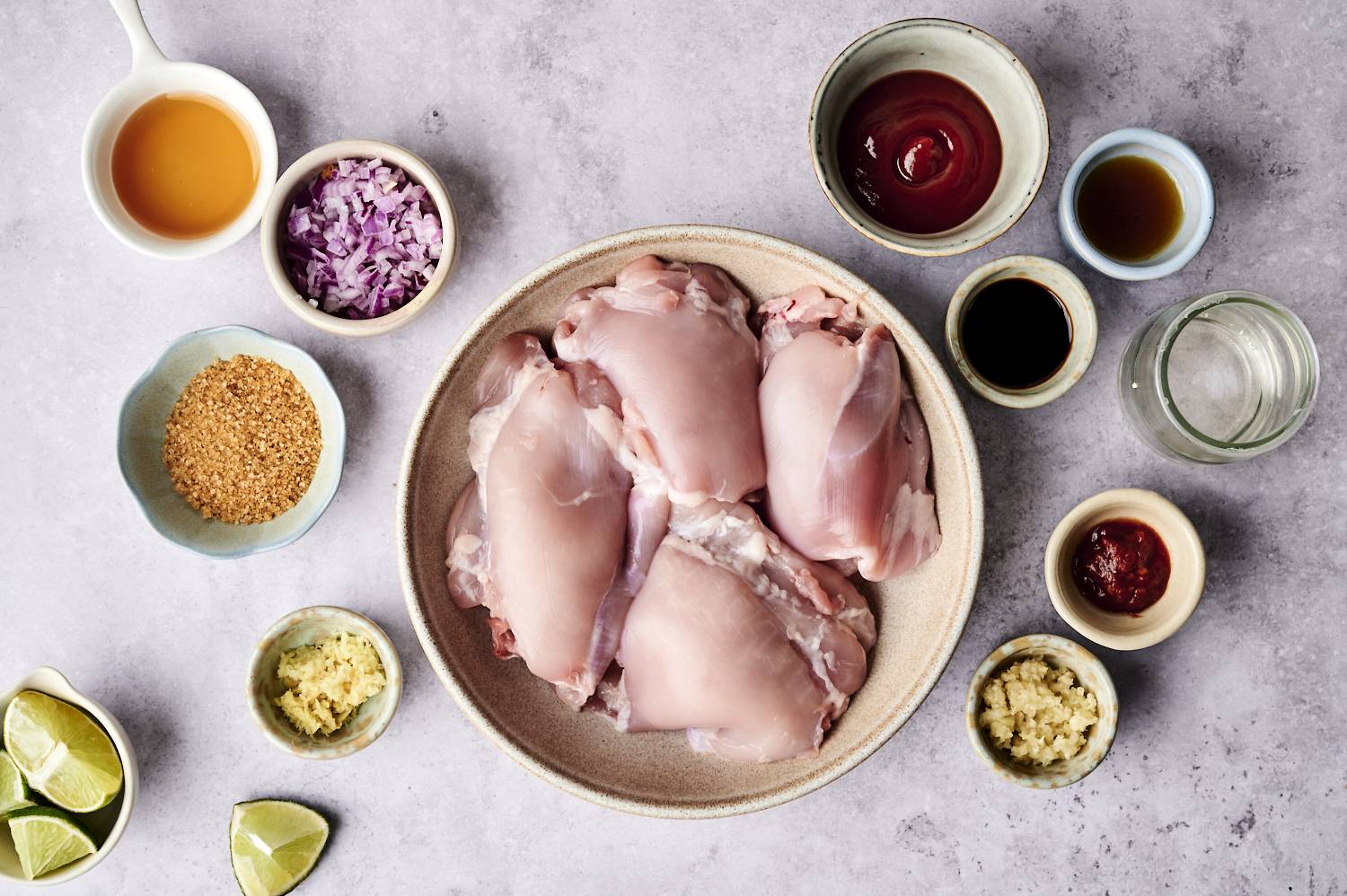 A bowl of raw chicken thighs surrounded by various ingredients, including chopped onions, sauces, spices, and lime wedges, on a countertop.