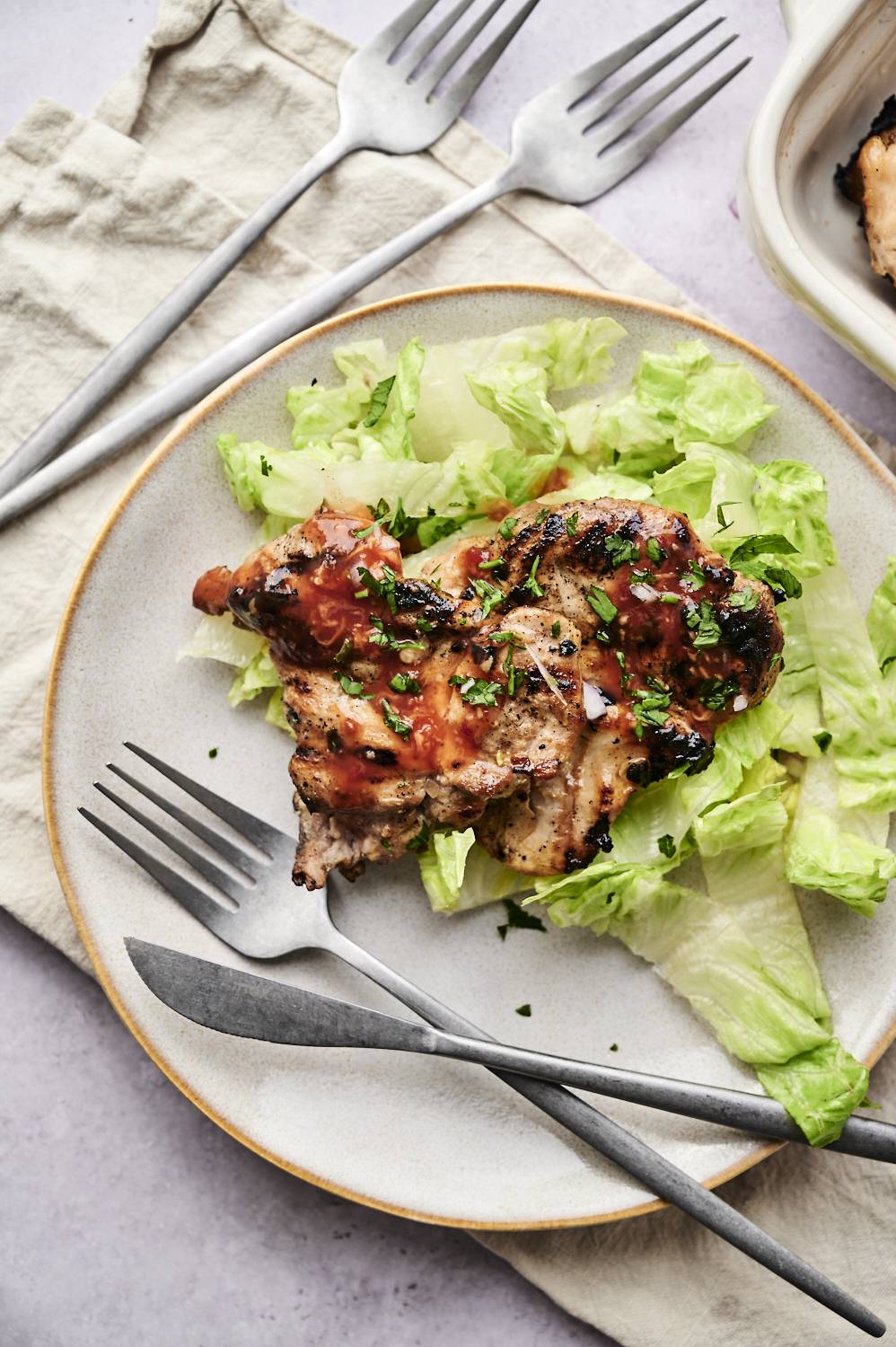 Grilled chicken thighs topped with sauce and herbs, served on a bed of lettuce, accompanied by silver cutlery on a textured table.
