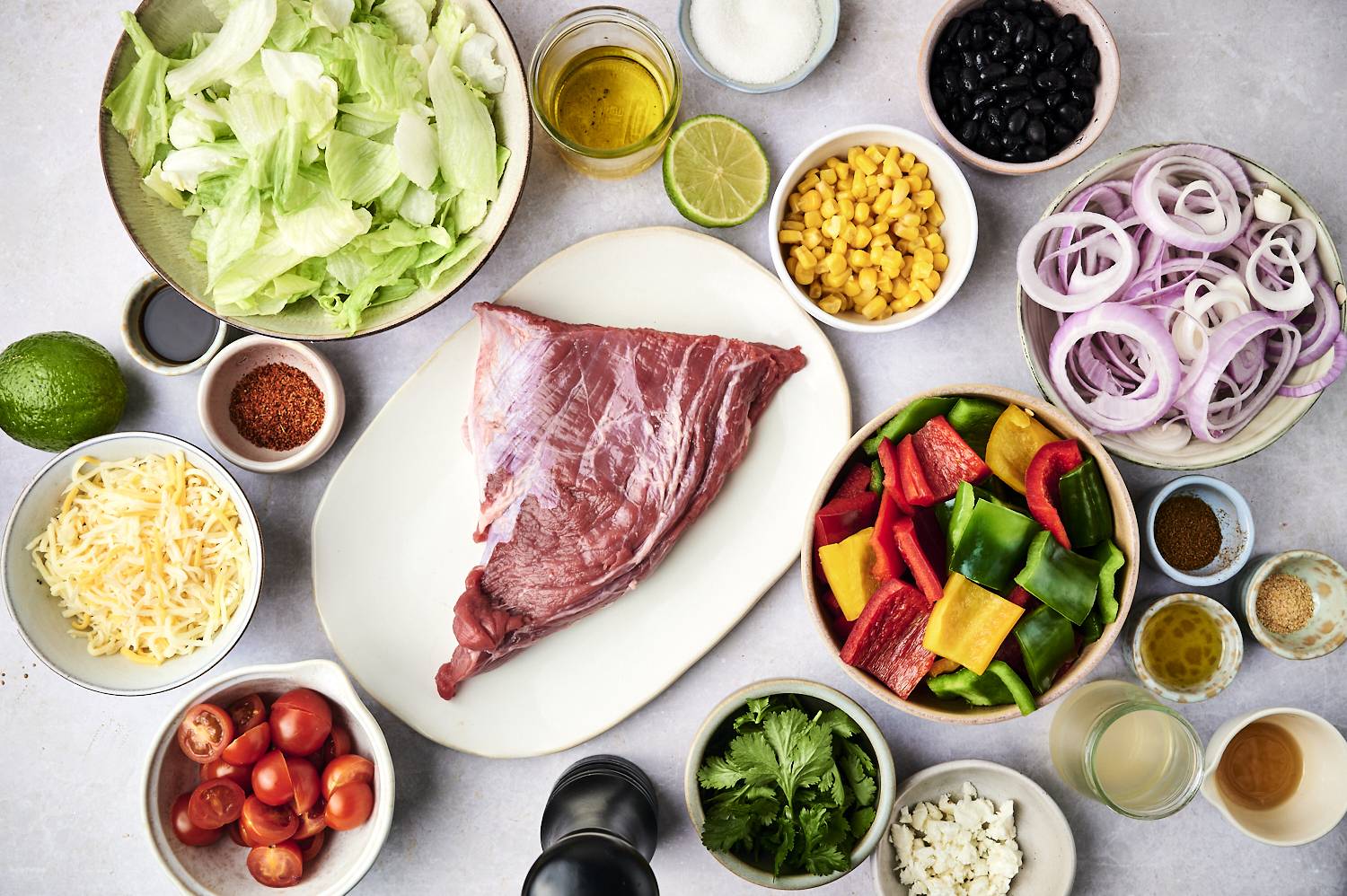 A wide variety of fresh ingredients for salads: lettuce, tomatoes, pepper, black beans, corn, cheese, onions and uncooked beef.