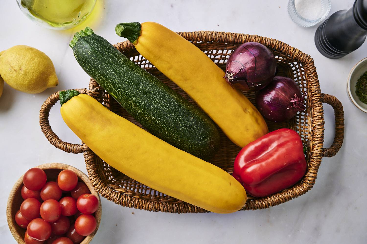 A woven basket filled with fresh zucchini, yellow squash, red bell pepper, and red onions, surrounded by cherry tomatoes and lemons.