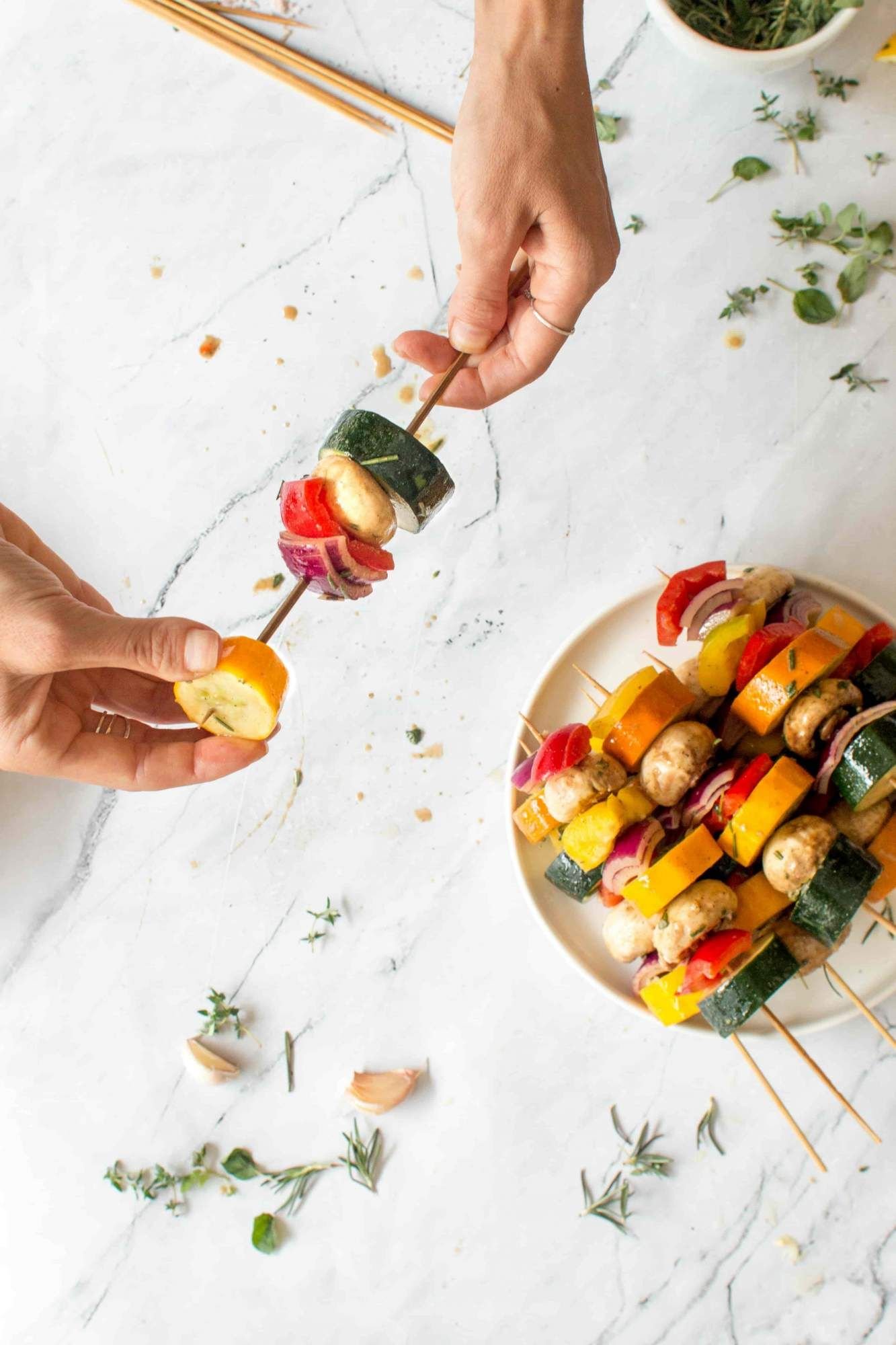 Vegetables being placed on a wooden skewer including zucchini, summer squash, red onion, pepper, and onion.