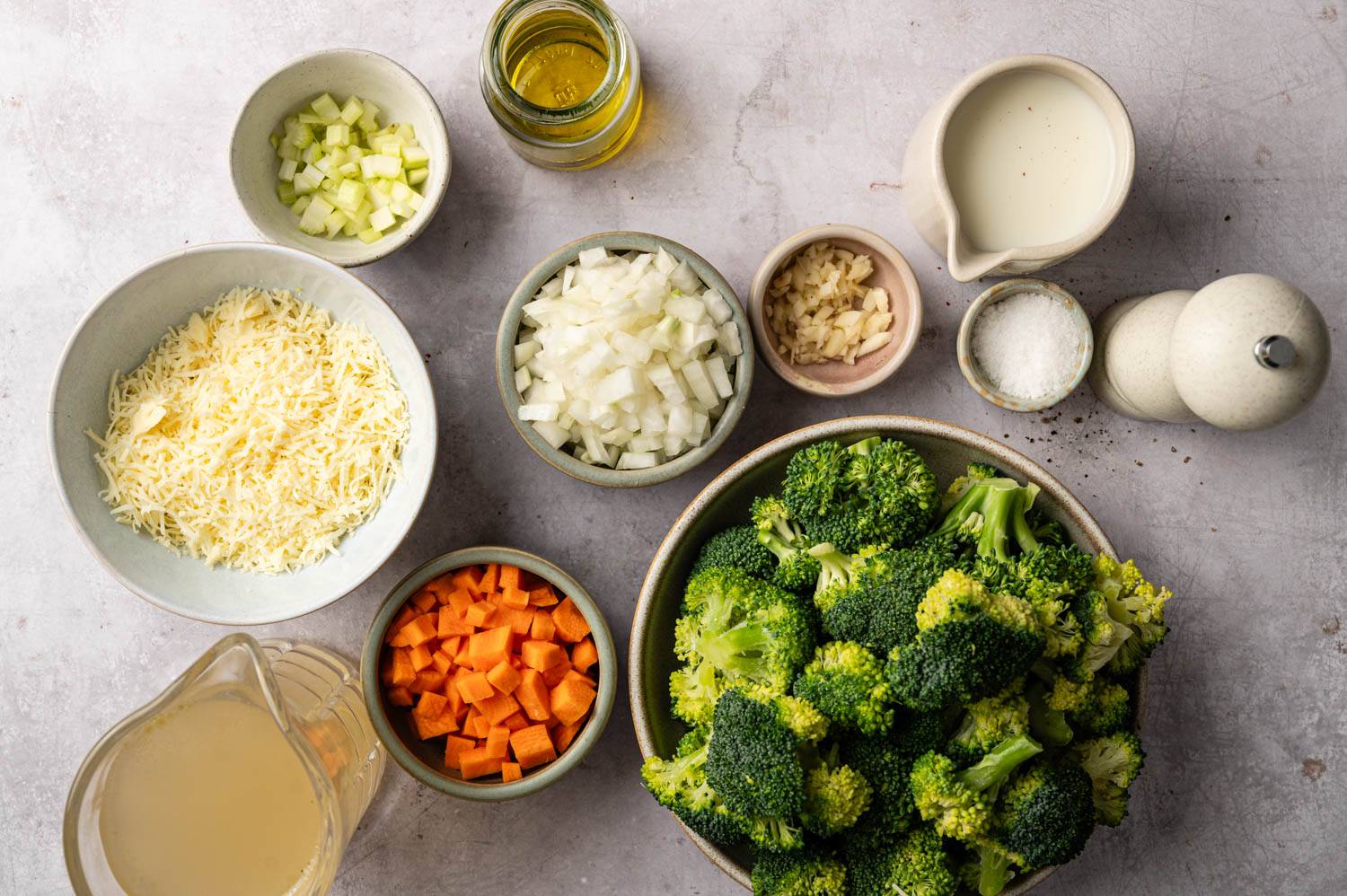 Assorted ingredients on a countertop for a broccoli soup recipe: chopped broccoli, onions, carrots, shredded cheese, garlic, cream, broth, oil, and seasonings.