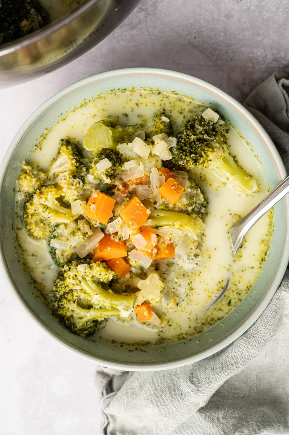 A bowl of creamy broccoli soup with visible broccoli florets, diced carrots, and onions. The soup appears warm and comforting, with a spoon resting inside. A light gray napkin is placed next to the bowl on a textured surface.