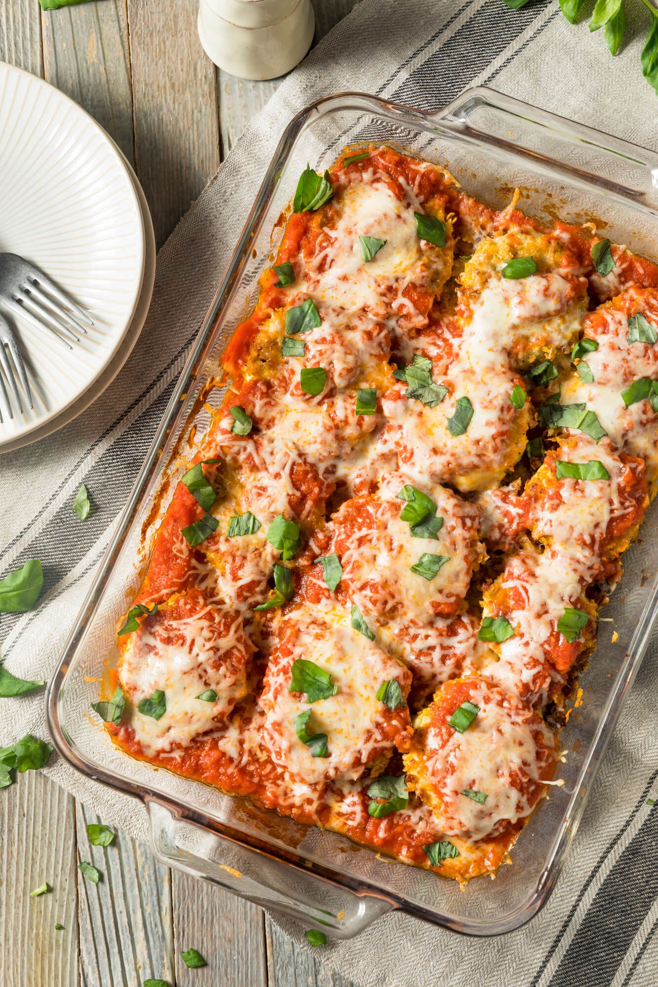 A wooden table with baked eggplant Parmesan in a glass casserole dish and basil.