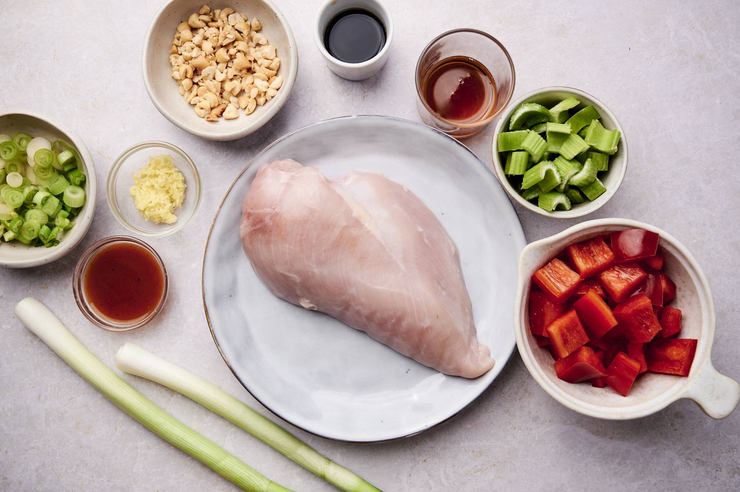 Ingredients for Healthy Kung Pao Chicken arranged neatly, including raw chicken breast, diced red bell peppers, sliced celery, chopped green onions, crushed peanuts, minced ginger, soy sauce, and various sauces in small bowls.