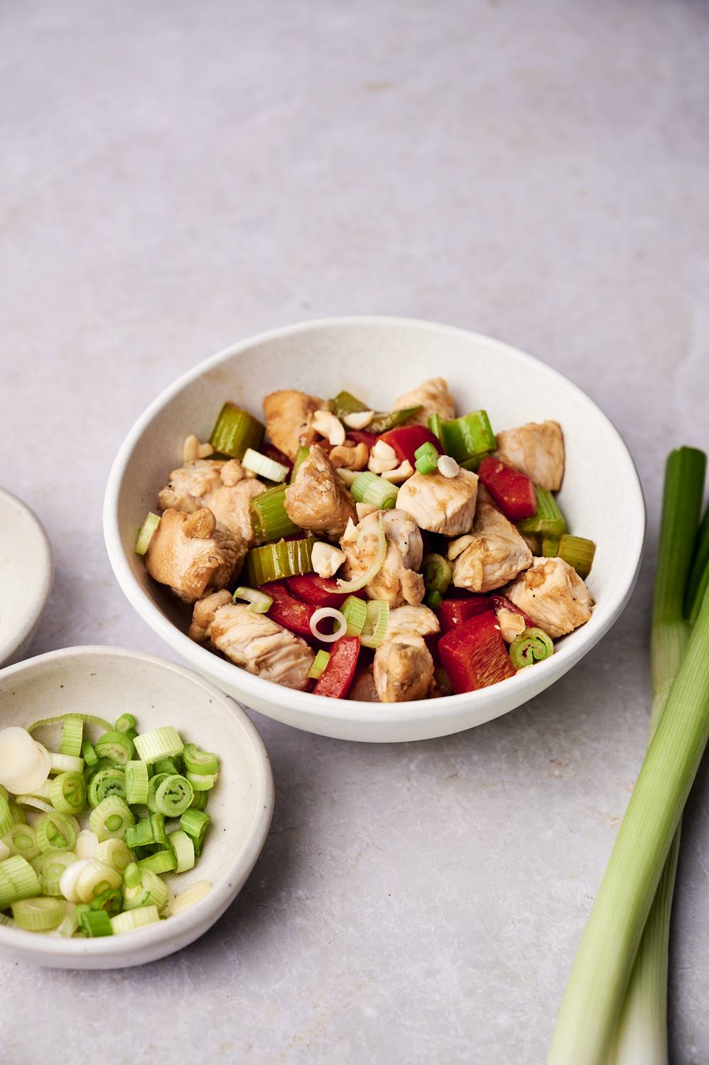 A bowl of Healthy Kung Pao Chicken featuring tender chicken pieces, red bell peppers, celery, and green onions, garnished with crushed peanuts. Fresh green onions and small dishes of sliced scallions are placed beside it.