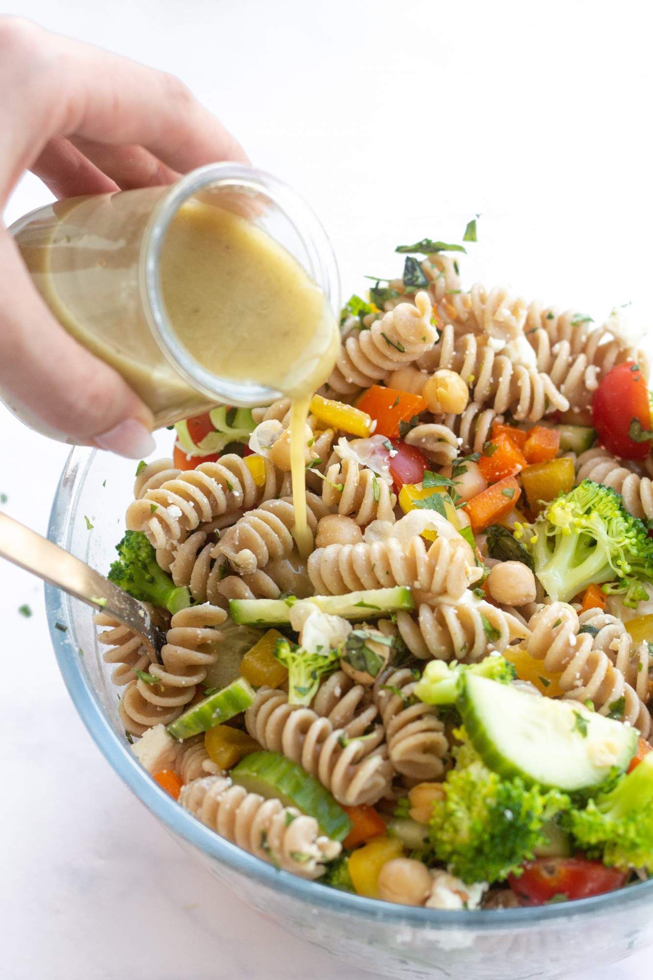 Whole wheat pasta salad in a bowl with cucumbers, broccoli, tomatoes, bell peppers, and feta cheese with dressing being poured on top.